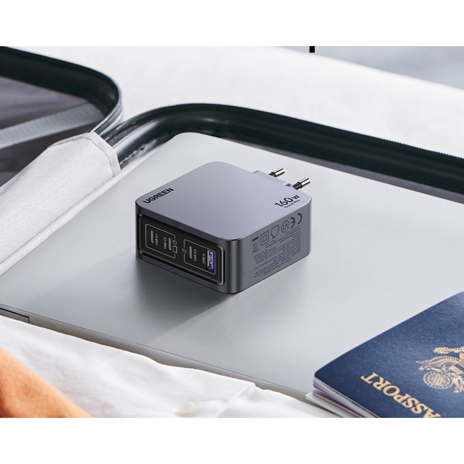 Caricatore USB-C grigio da 160W su laptop accanto a passaporto in valigia aperta.