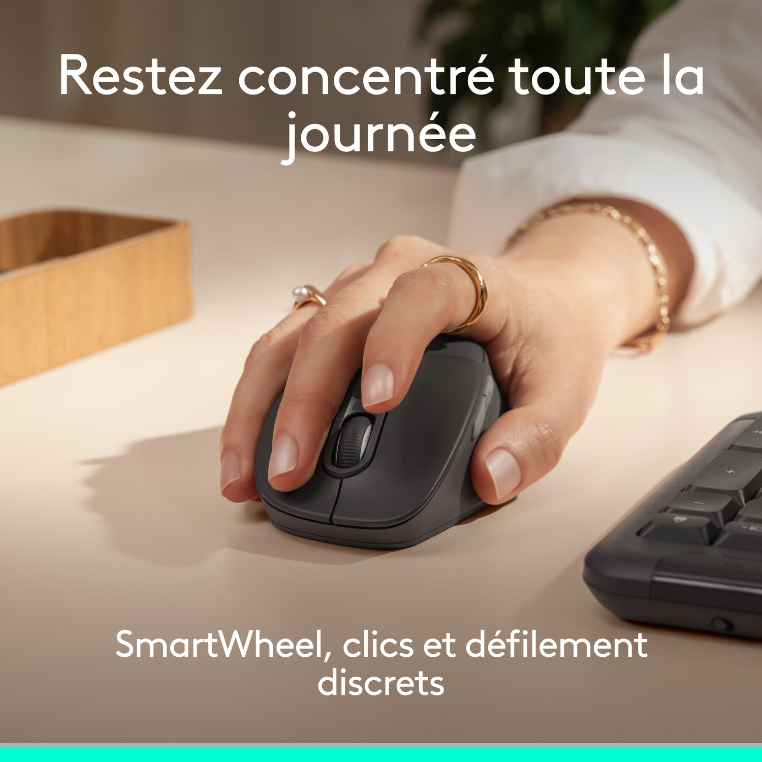 Une main utilise une souris grise à côté d'un clavier sur un bureau brun clair.