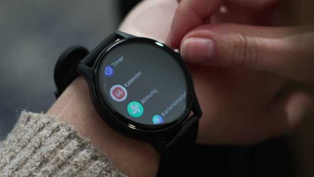 Eine Person bedient eine Smartwatch mit rundem Display, auf dem Apps wie Timer und Kalender angezeigt werden.