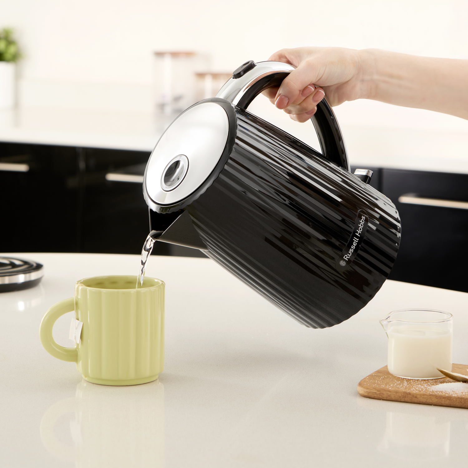 A hand pours hot water from a black kettle into a light green mug with a tea bag.