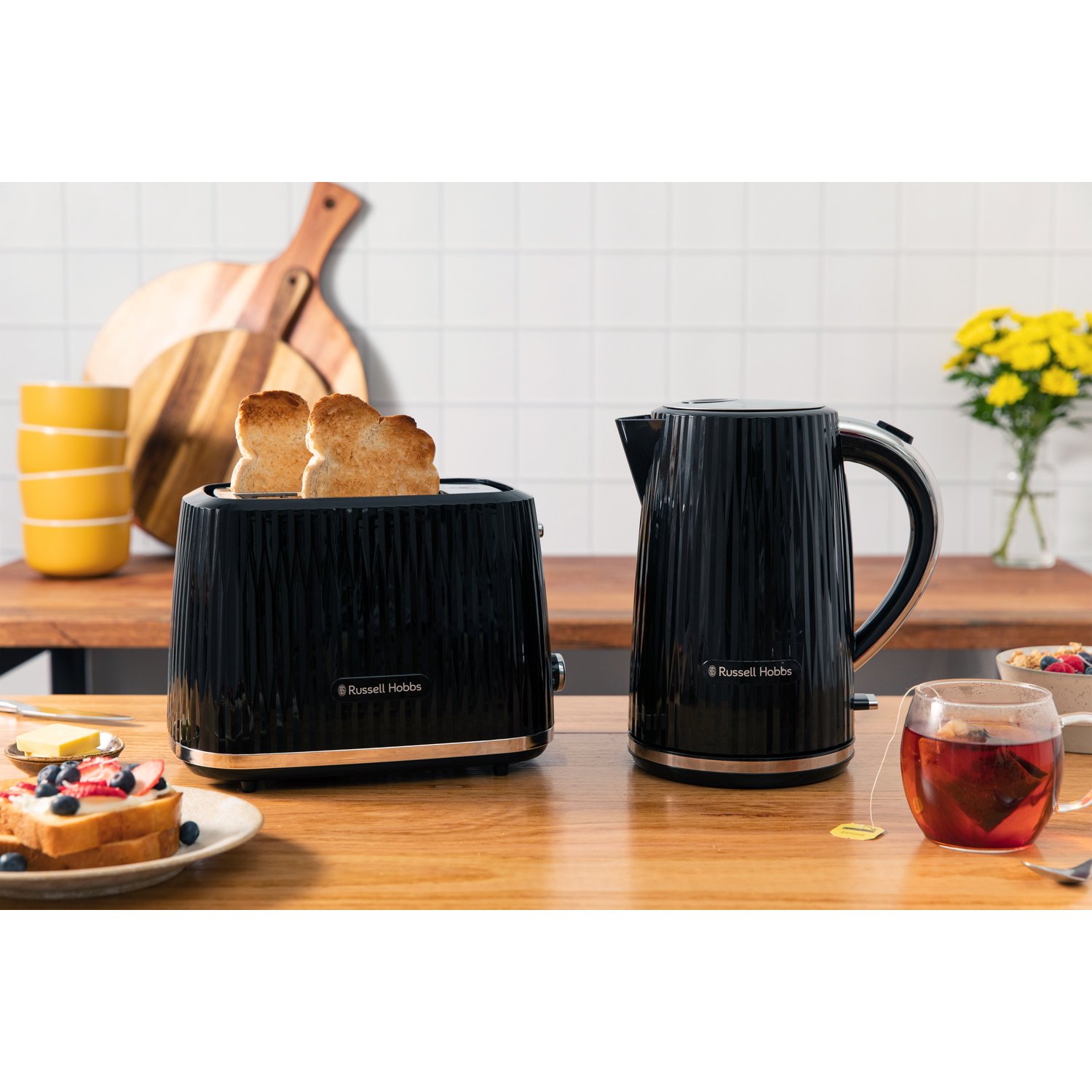 Black toaster with toast, kettle, tea, and pastries on a wooden table.