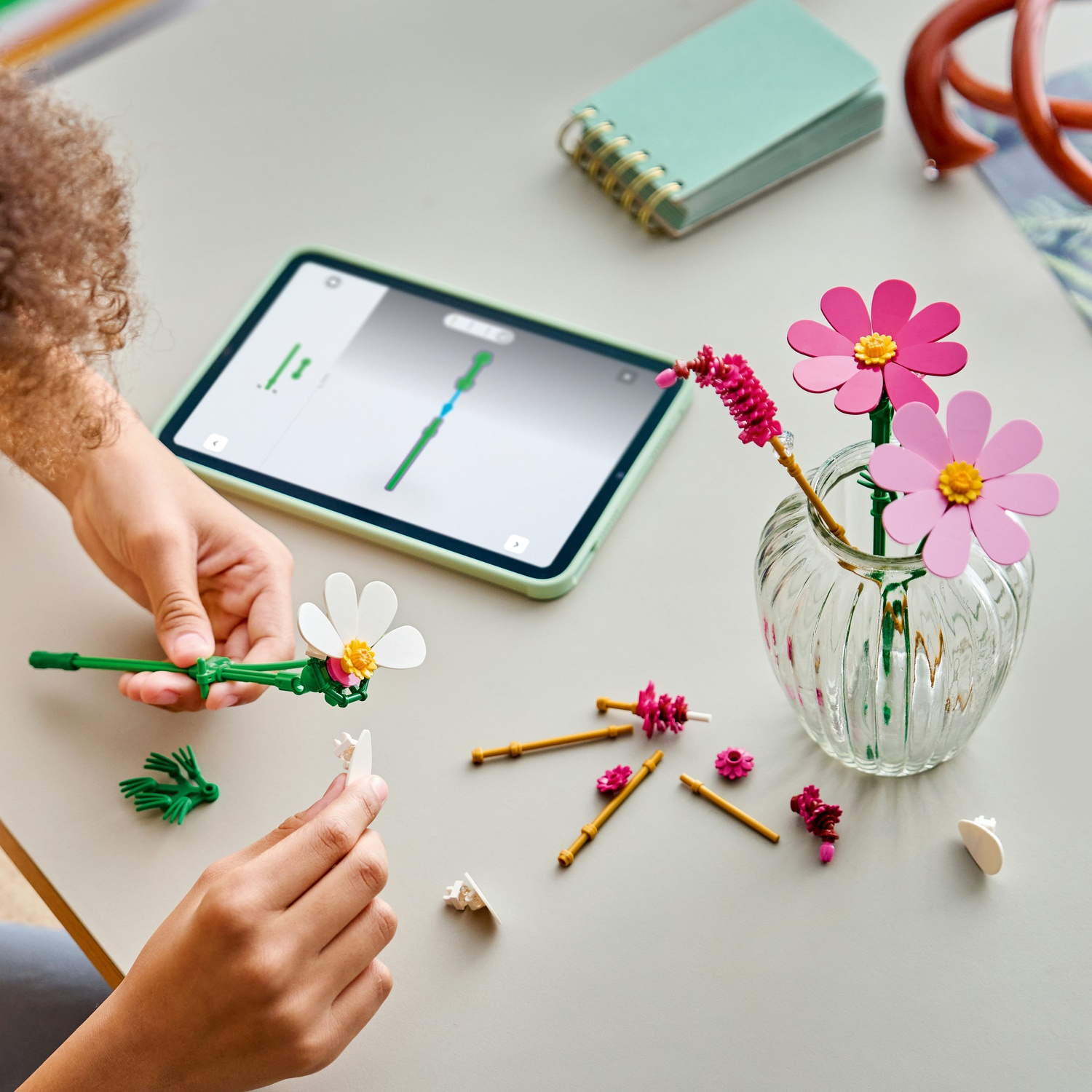 Mani che assemblano un fiore bianco; un vaso con fiori rosa e un tablet sono sul tavolo.