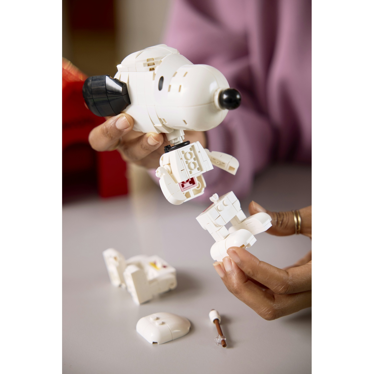 Mani assemblano un modello Lego bianco di Snoopy. Pezzi e attrezzo sul tavolo.