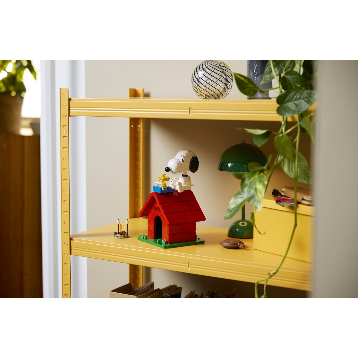 Snoopy siede sopra la sua cuccia rossa su uno scaffale giallo con piante e decorazioni.