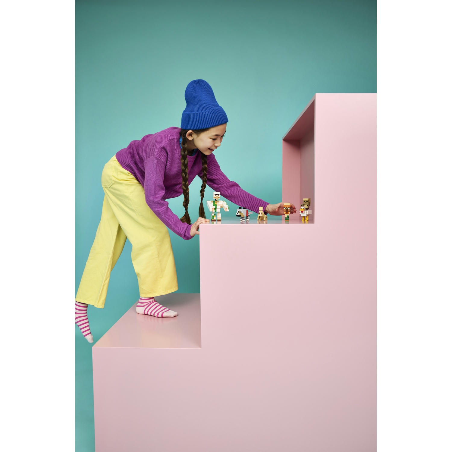 Una ragazza con cappello blu e maglione viola sistema figure su gradini rosa.
