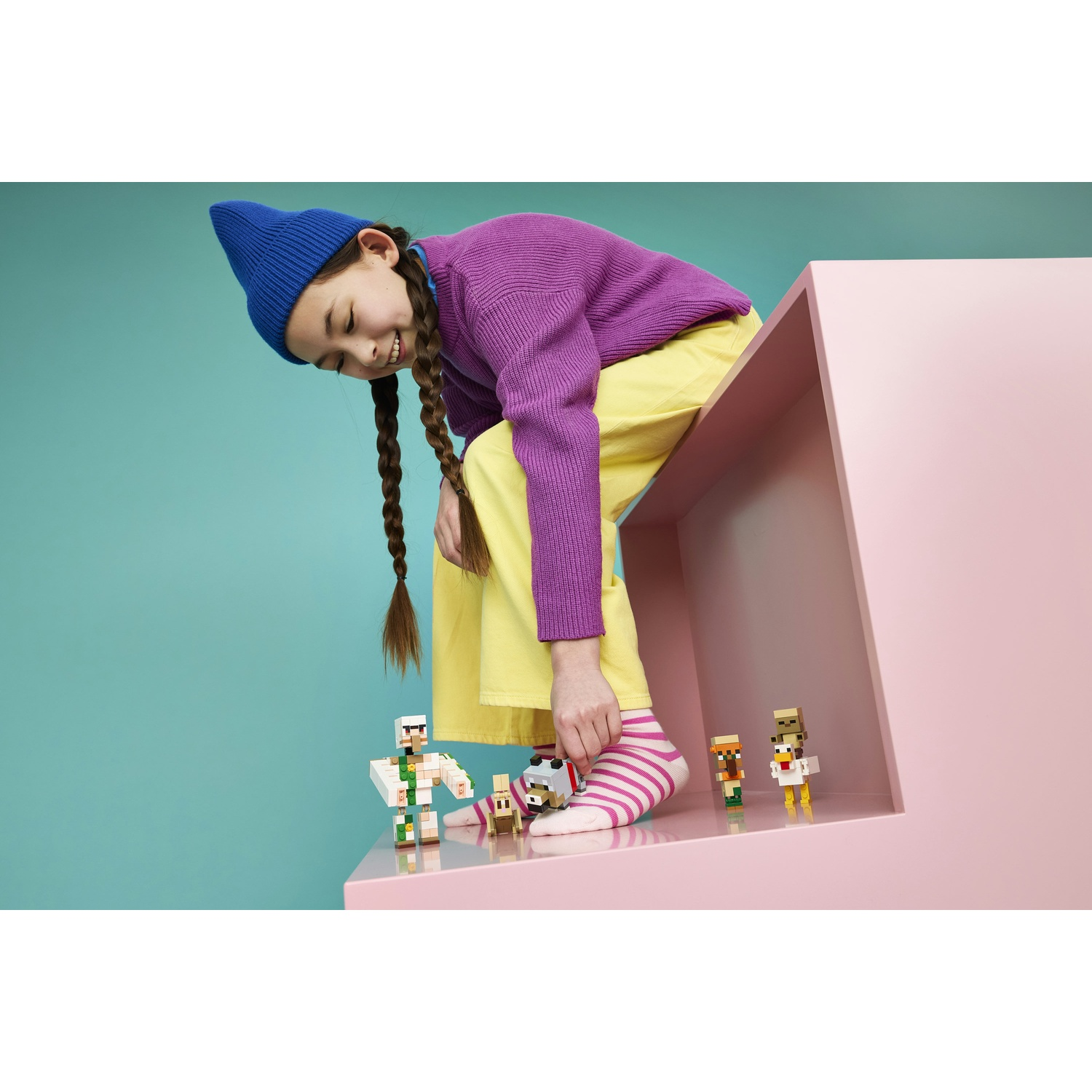 Bambina con cappello blu e maglione viola gioca con figure Minecraft su mensola rosa.