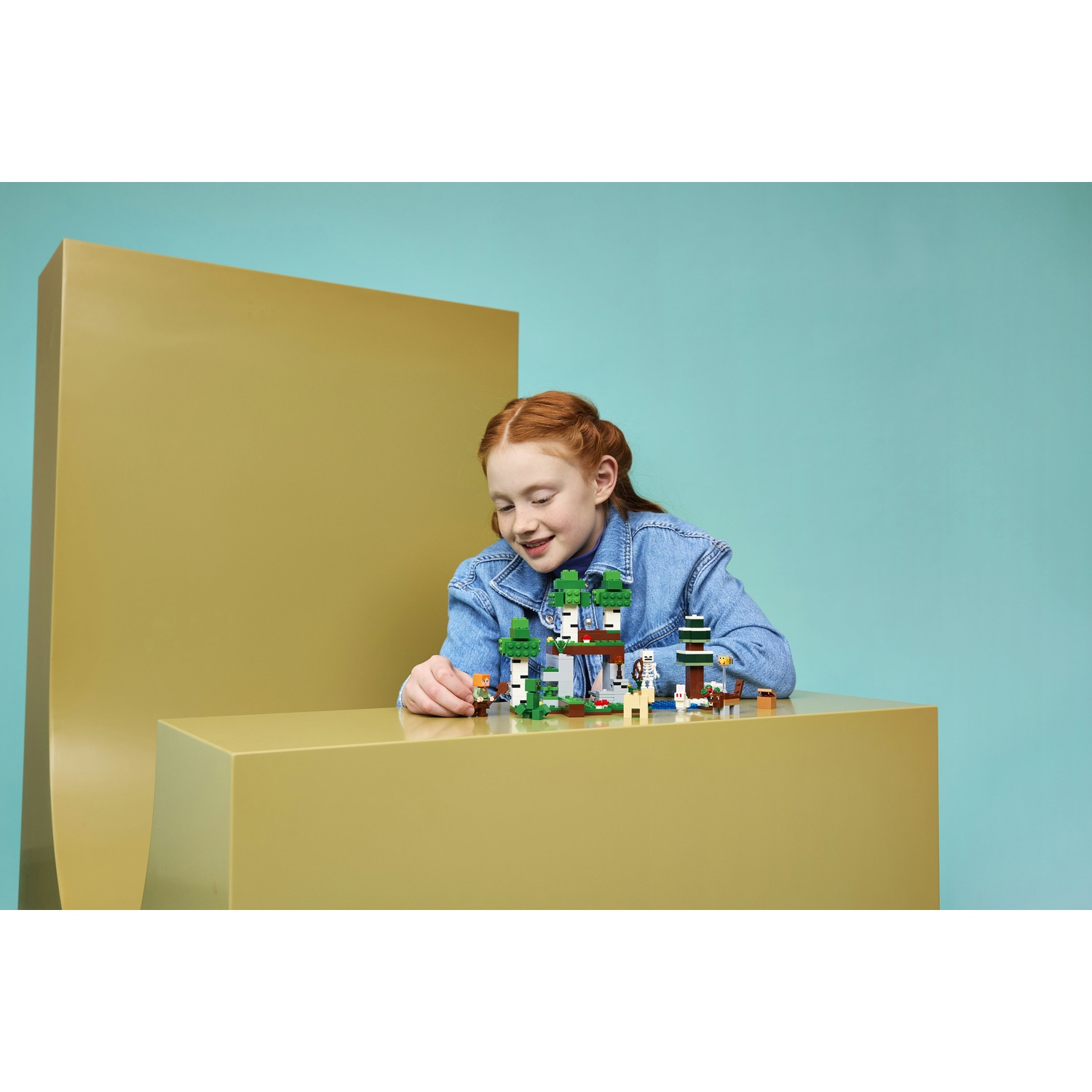Una ragazza sorride, giocando con un set Lego Minecraft con alberi e figure.