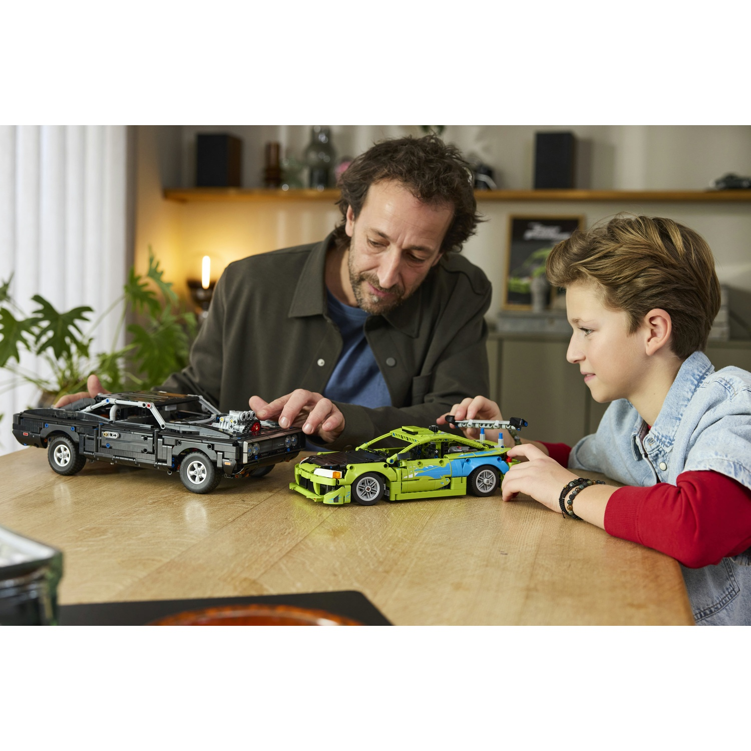 Uomo e ragazzo costruiscono modelli Lego di due auto su tavolo di legno.