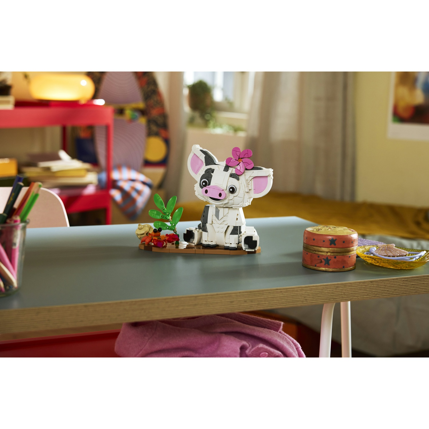 Un giocattolo Lego di Heihei siede su una scrivania, con un fiore rosa sulla testa. Una piccola pianta e un granchio accanto.