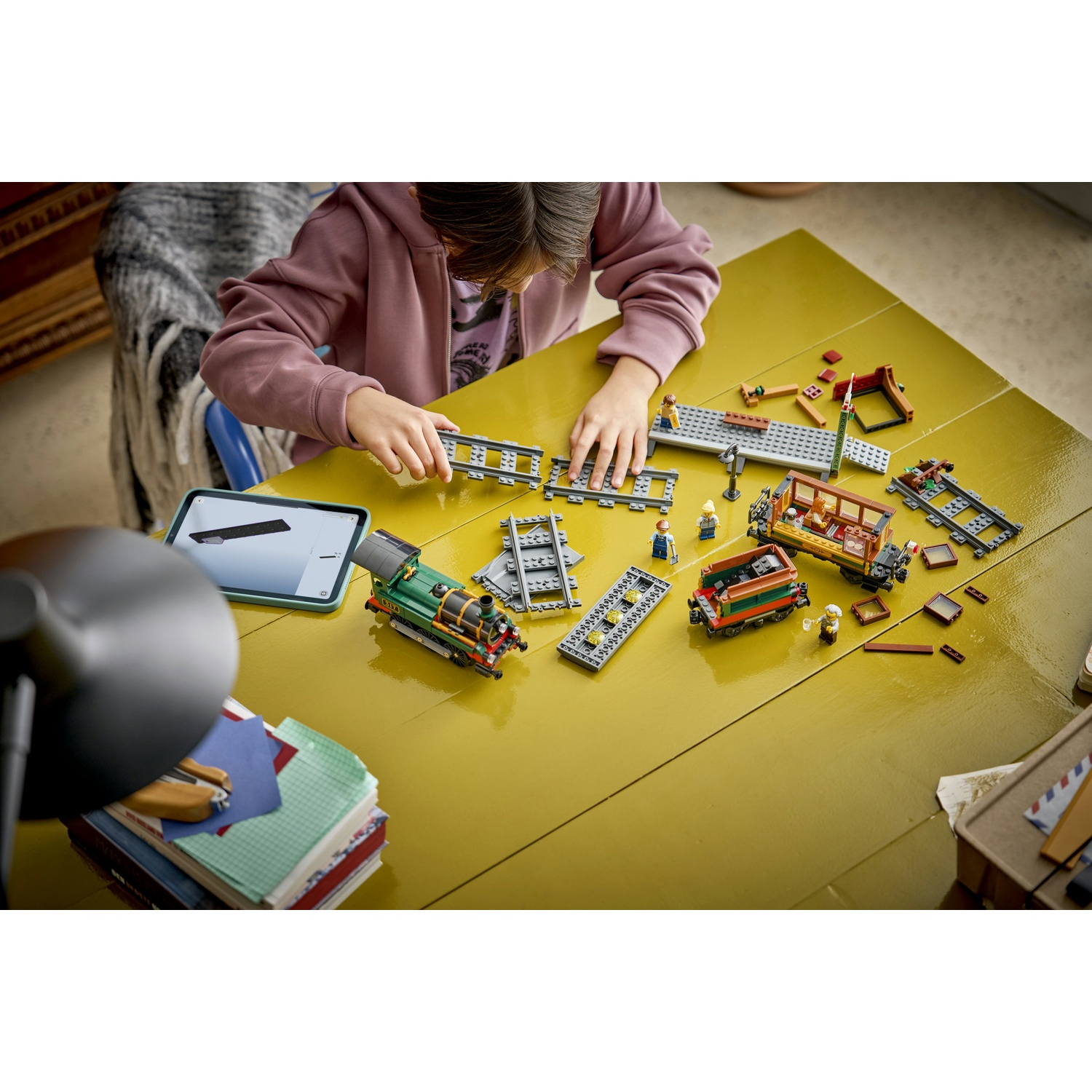 Un bambino costruisce un trenino Lego su un tavolo giallo, con treni, binari e figure.