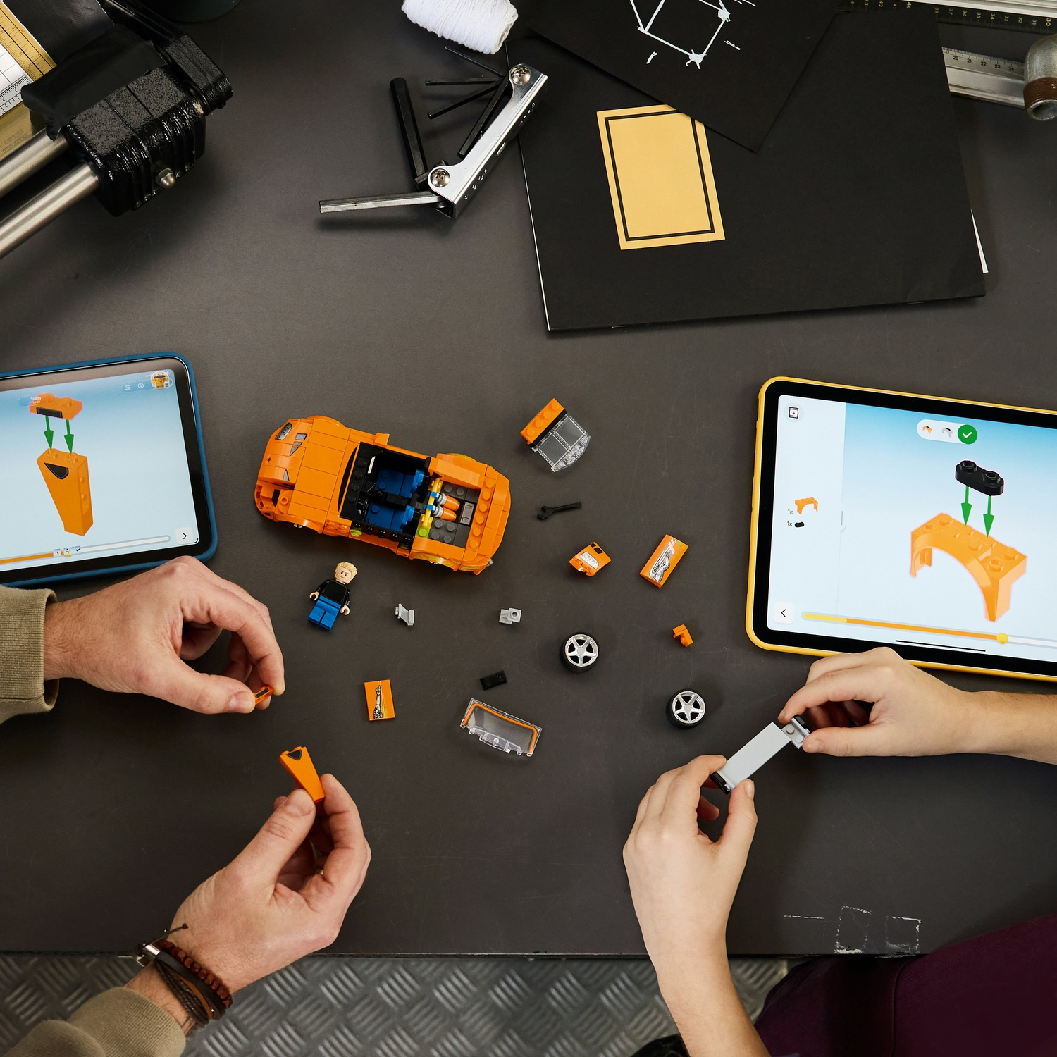 Mani assemblano pezzi di auto Lego arancioni su tavolo scuro con tablet che mostrano istruzioni.