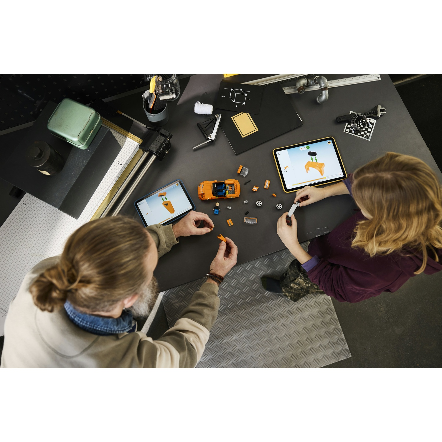 Due persone costruiscono un'auto Lego arancione, guardando tablet.