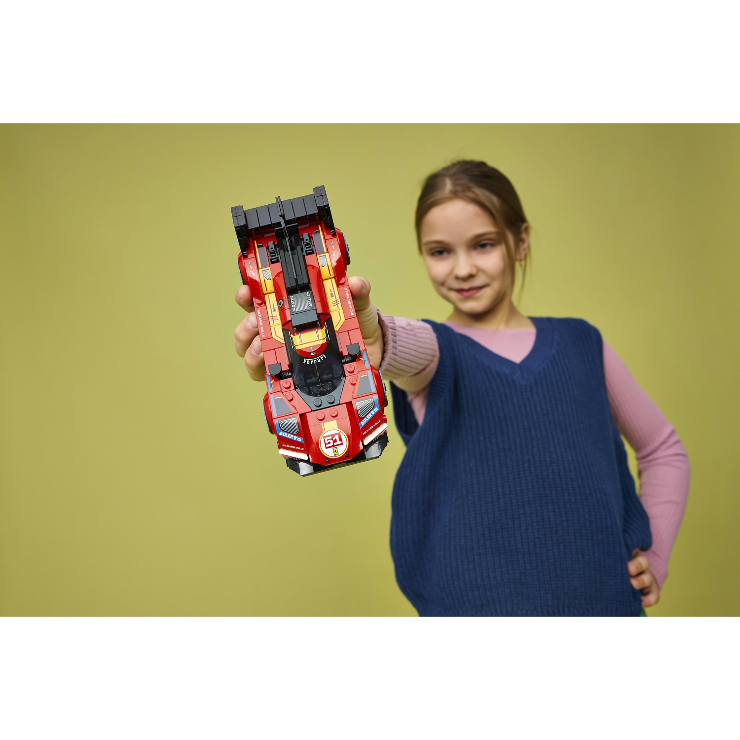 Una bambina tiene in mano un'auto da corsa Lego Ferrari rossa numero 51. Indossa un gilet blu.