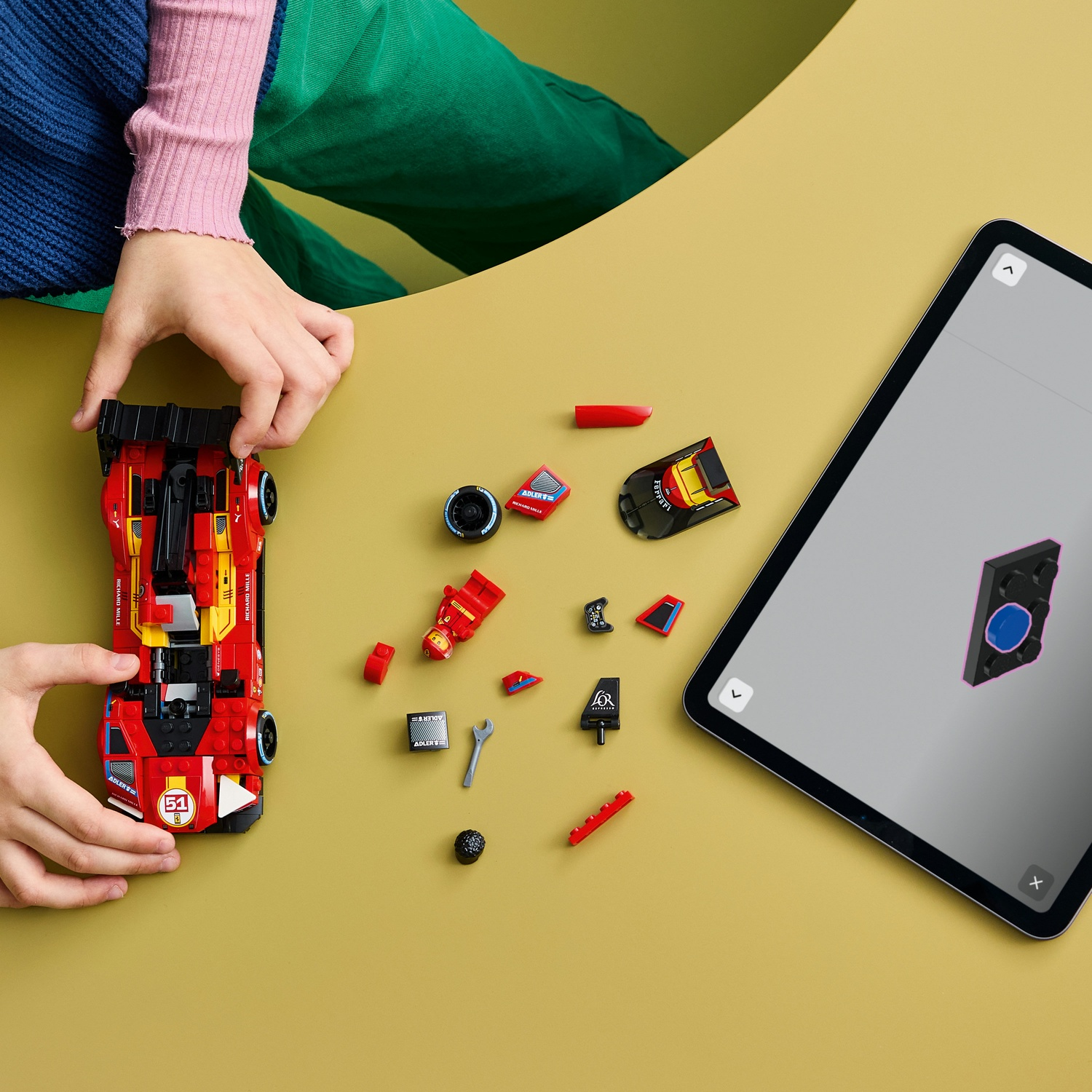 Mani assemblano un'auto LEGO rossa accanto a pezzi sparsi e un tablet che mostra le istruzioni.