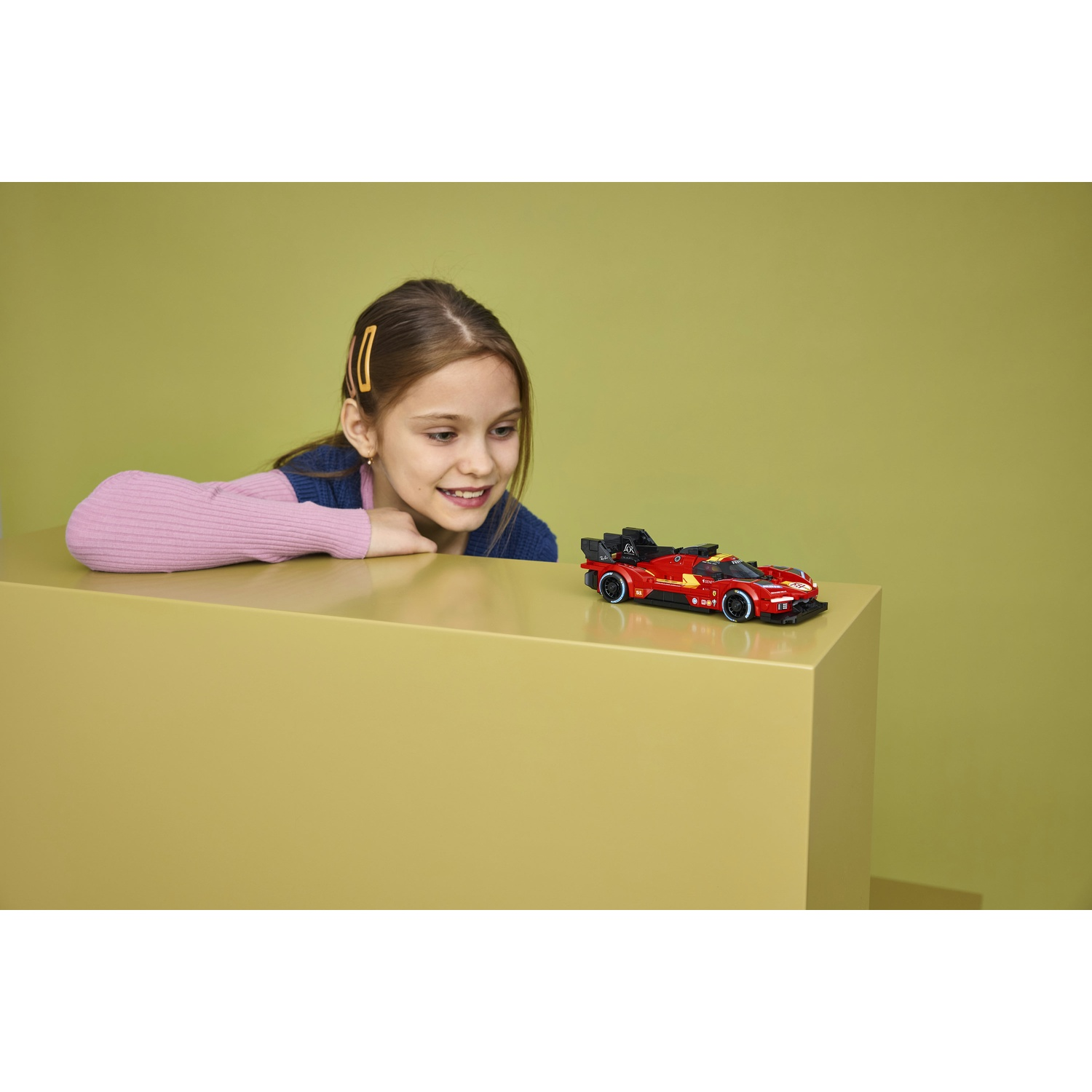 Una bambina guarda una macchinina da corsa rossa su un cubo giallo.