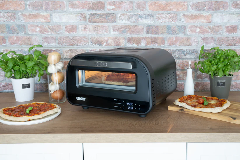 Un four à pizza Unold noir avec une pizza à l'intérieur. Des pizzas sont placées à gauche et à droite.