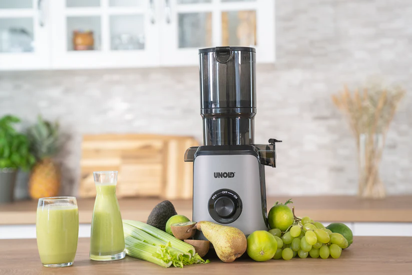 Ein Entsafter steht auf einem Holztisch mit Obst und Gemüse wie Äpfeln, Birnen, Kiwi, Avocado, Limetten, Trauben, Sellerie und einem Glas Saft.