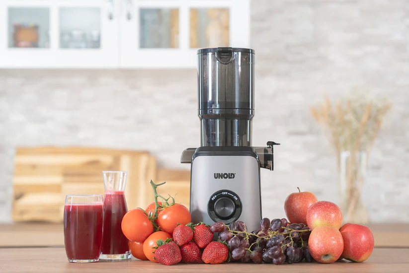 Ein Entsafter steht neben Obst und Gemüse, mit zwei Gläsern Saft.