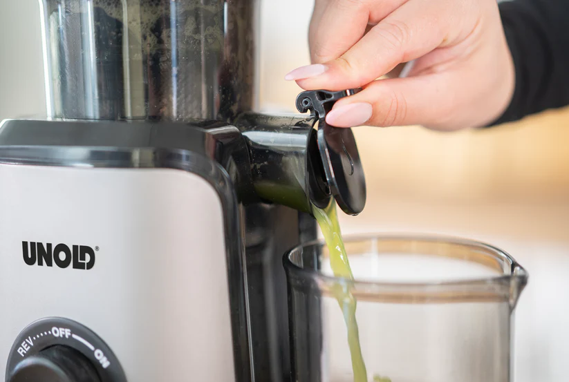 Grüner Saft fließt aus einem Entsafter in einen Behälter. Eine Hand öffnet den Auslauf.