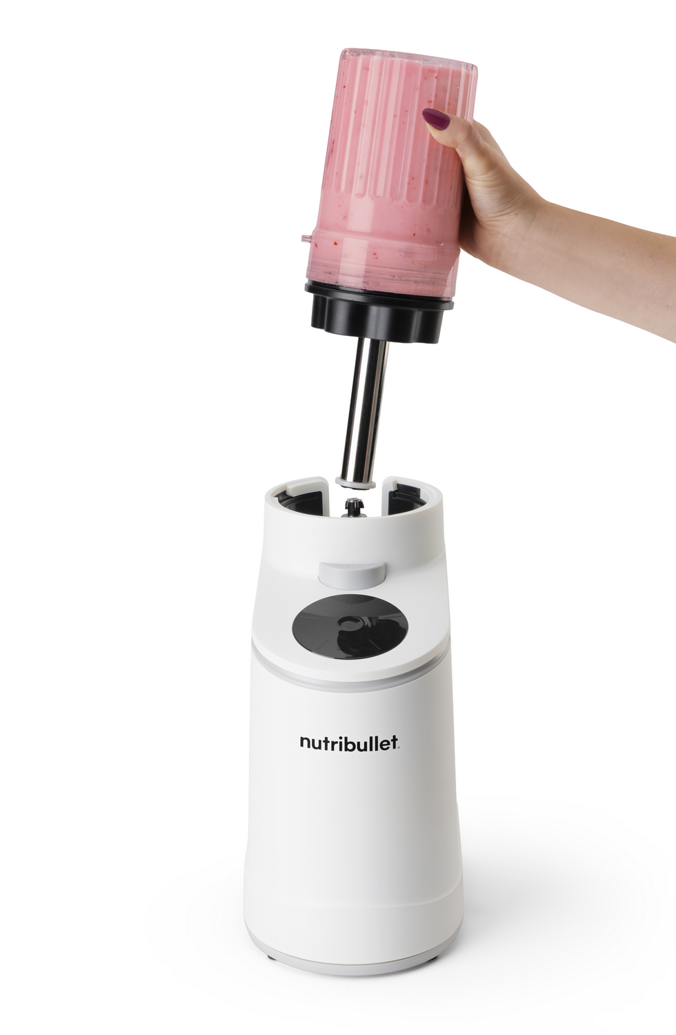 Una mano sostiene una taza con batido rosa que se coloca en una licuadora blanca.