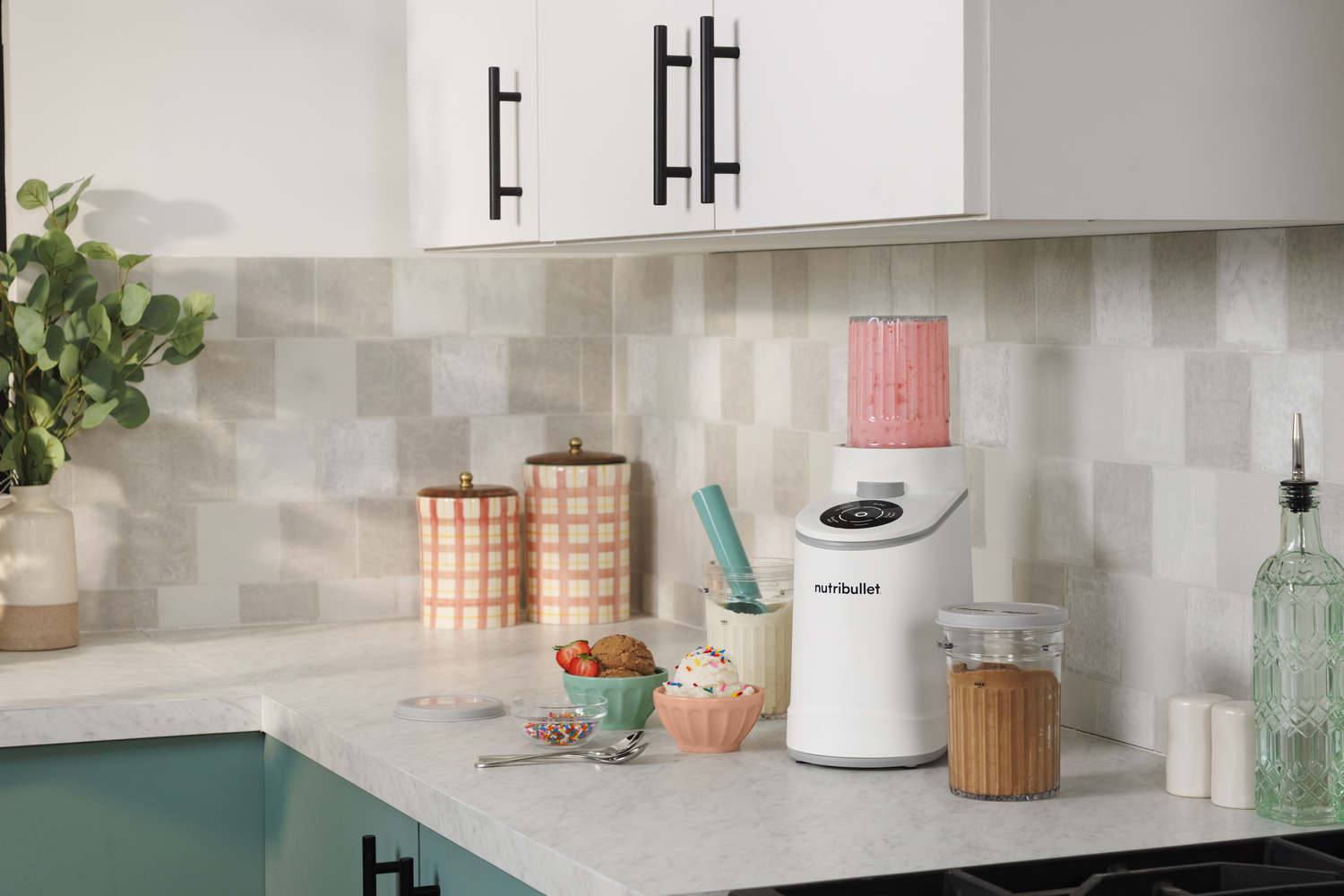 Una licuadora Nutribullet blanca con batido rosa está sobre un mostrador con helado, chispas y fresas.