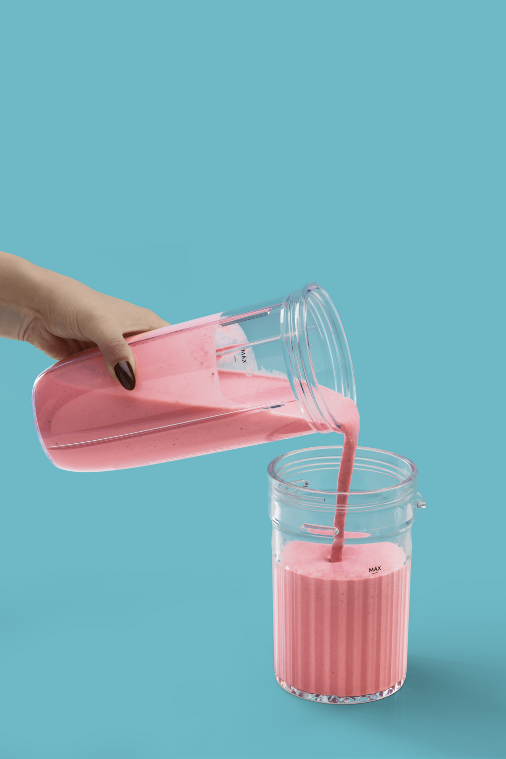 Una mano vierte batido rosa de un vaso transparente a otro vaso transparente sobre fondo azul.