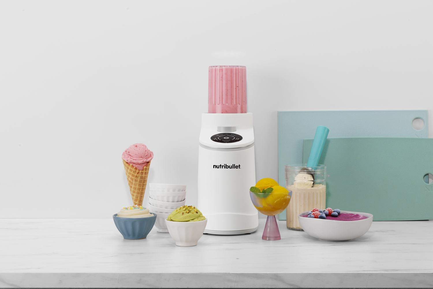 Una licuadora Nutribullet blanca sobre un mostrador de mármol rodeada de helados y cuencos de fruta.