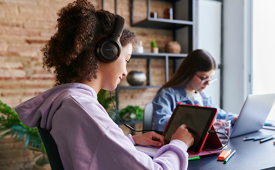 Eine junge Person mit Locken und Kopfhörern benutzt ein Tablet, während eine andere Person im Hintergrund an einem Laptop arbeitet.