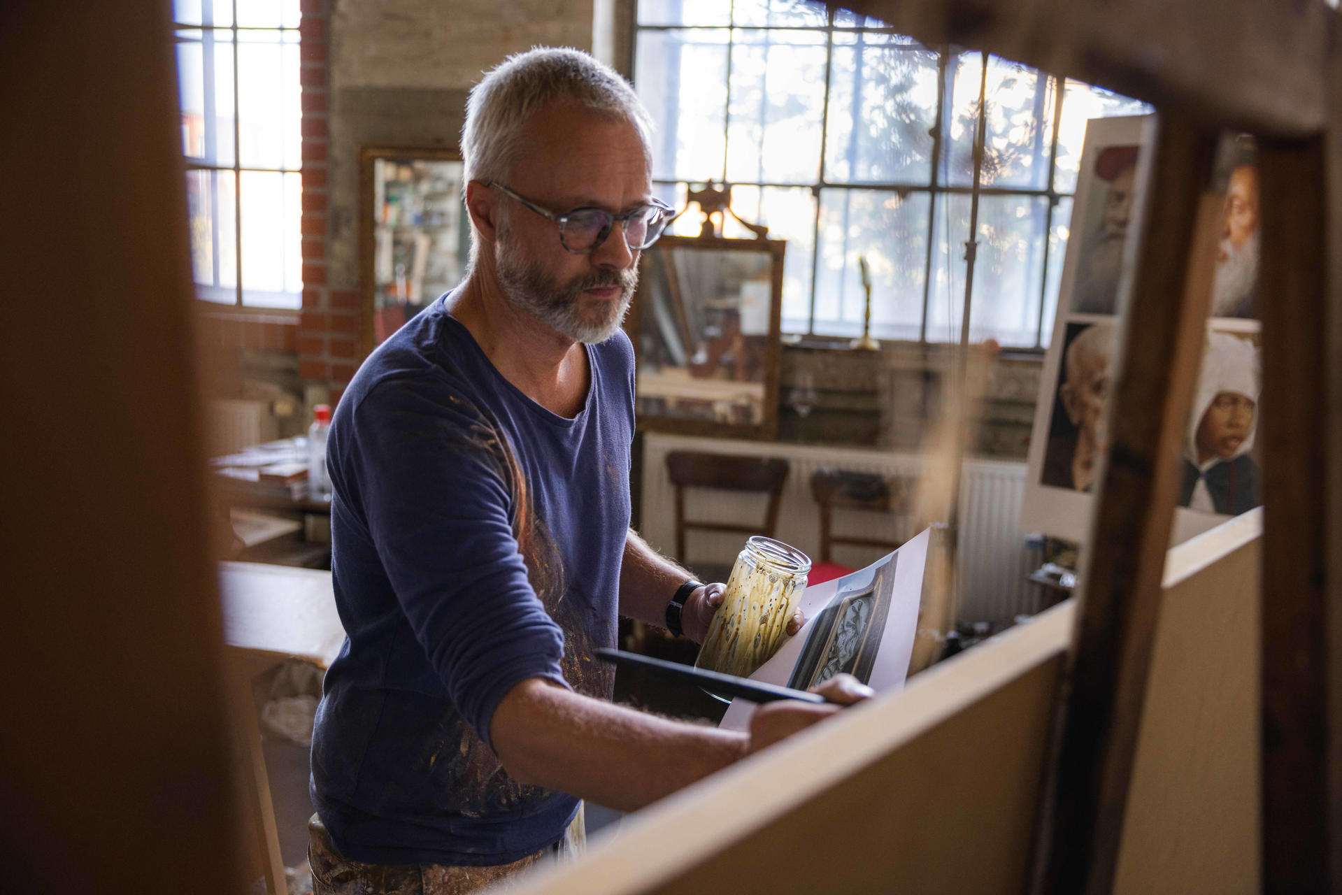 Ein Mann mit Brille und Bart malt in einem Atelier. Er hält eine Farbflasche und einen Pinsel.