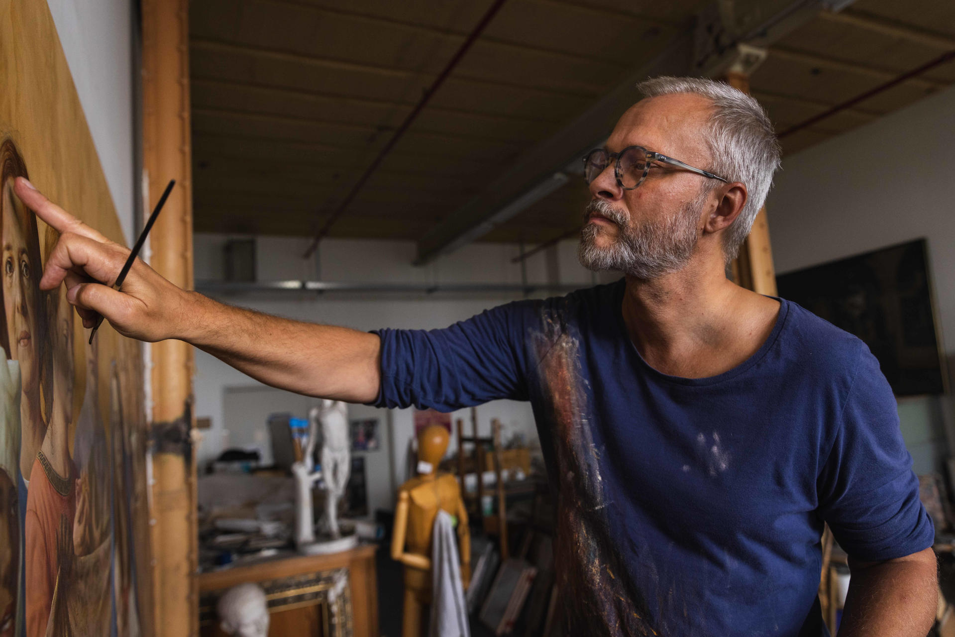 Ein Mann mit Brille und grauem Bart malt ein Gemälde in einem Atelier.