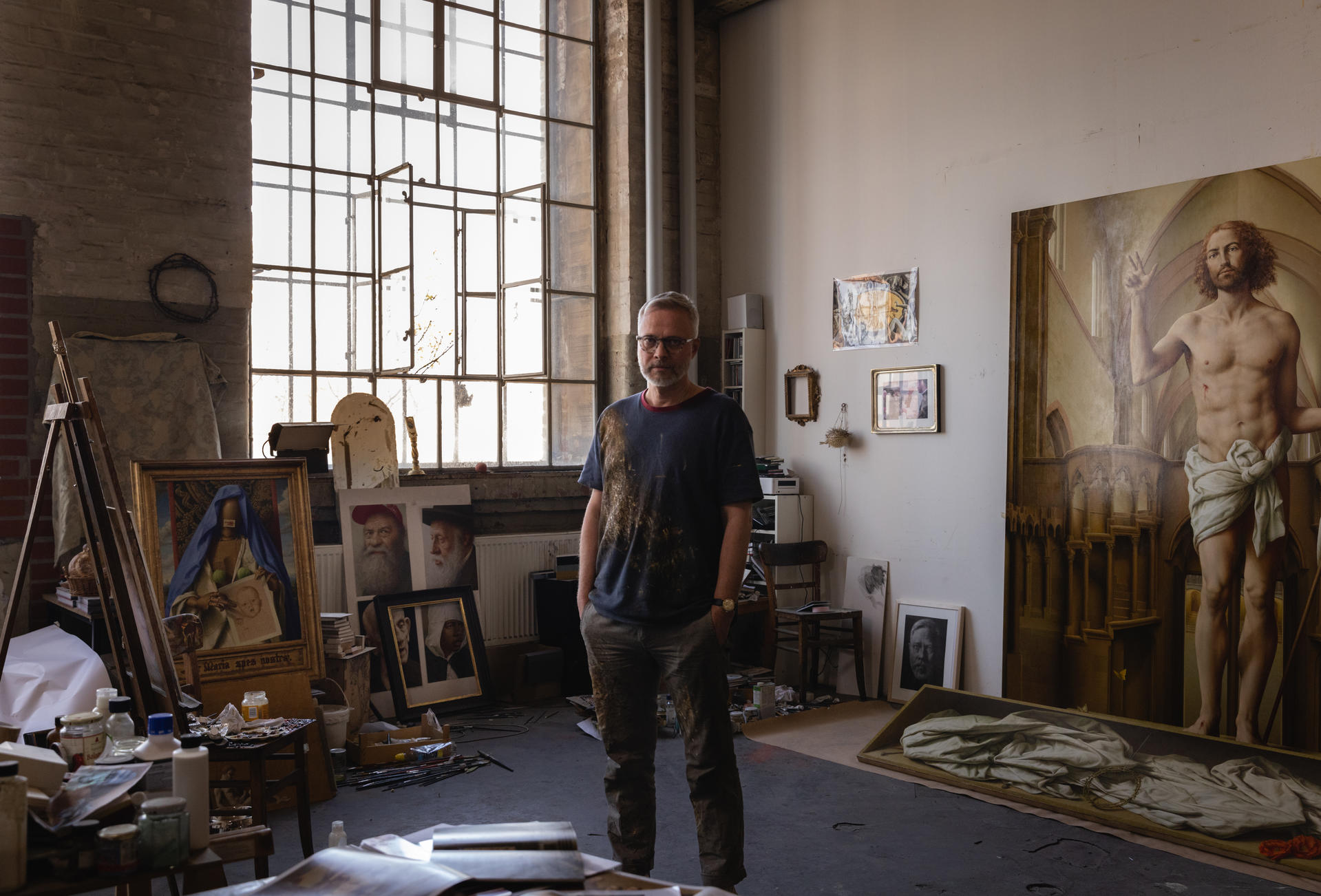Künstler im Atelier mit Gemälden, großem Fenster, Staffelei und Kunstbedarf.