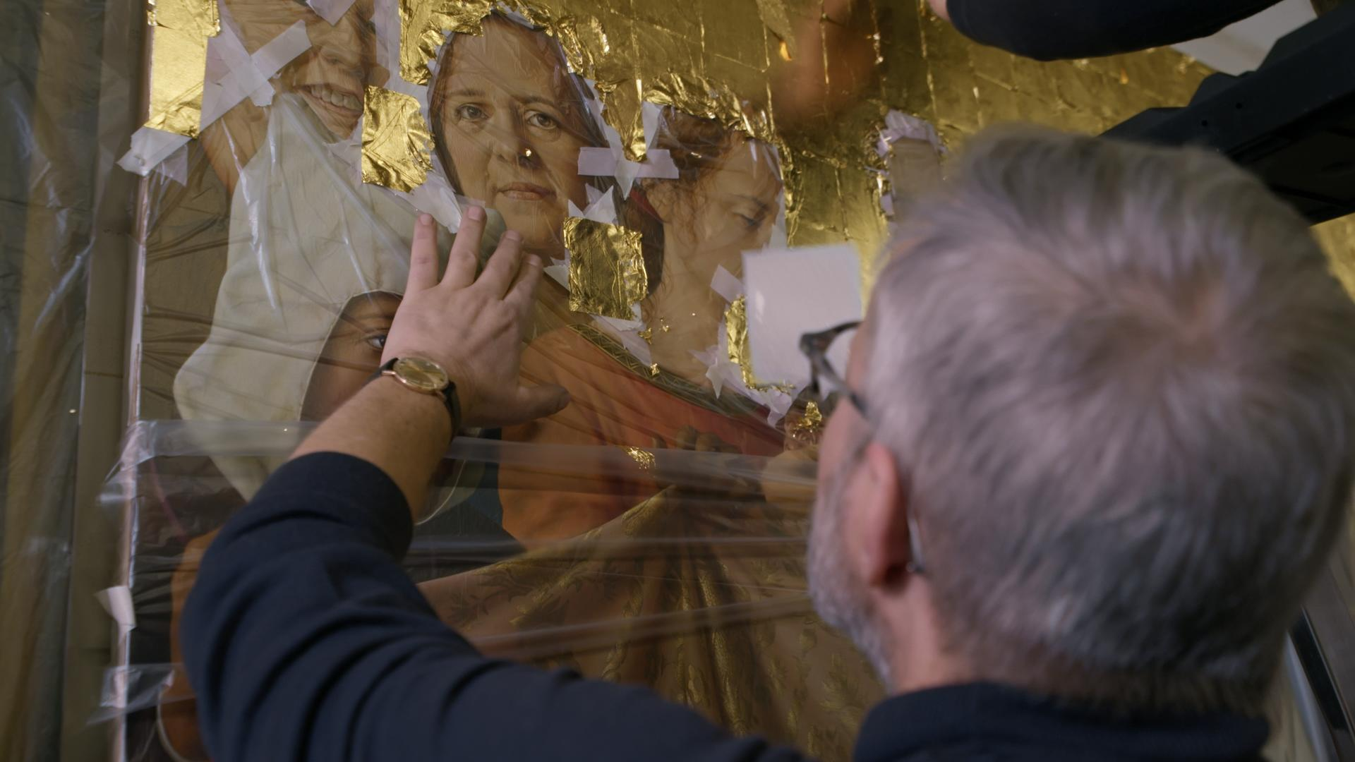 Mann mit Brille und grauem Haar berührt eine goldene, schimmernde Kunst mit Porträts.