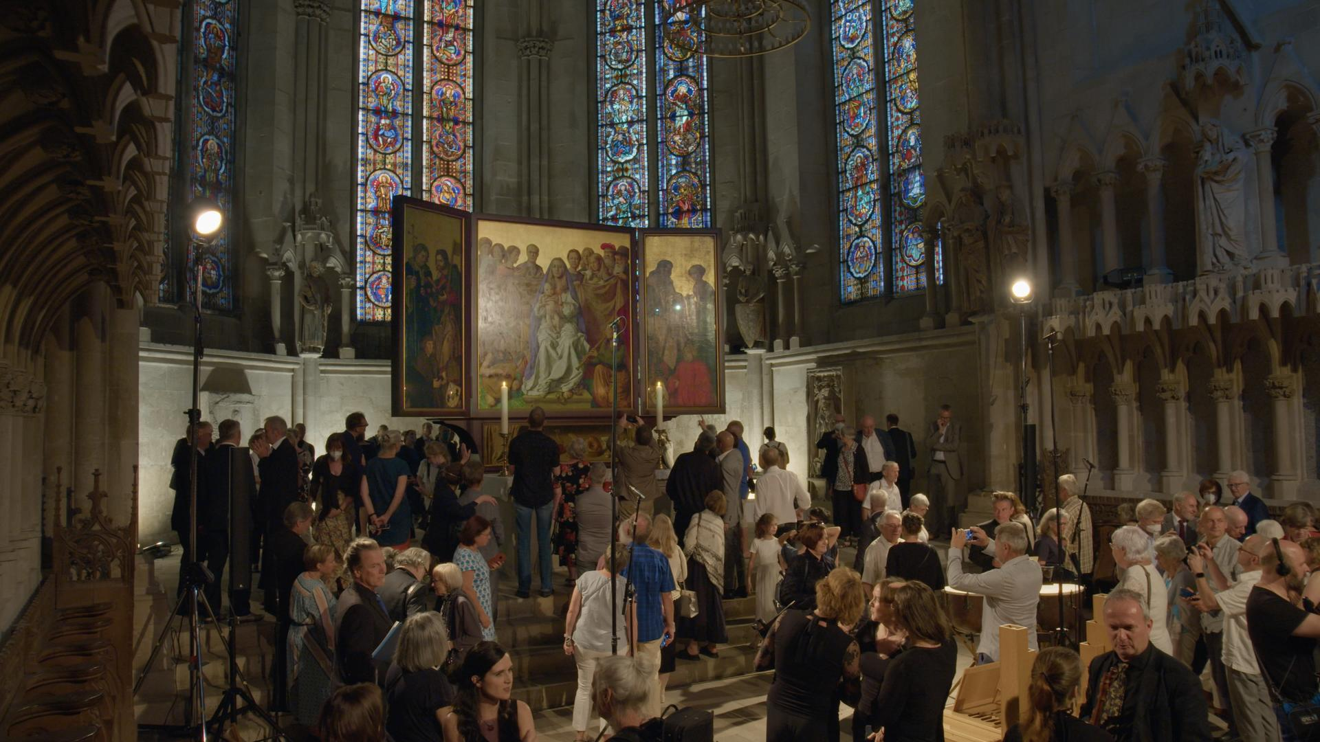 Menschen in einer Kirche vor einem Triptychon und bunten Buntglasfenstern.