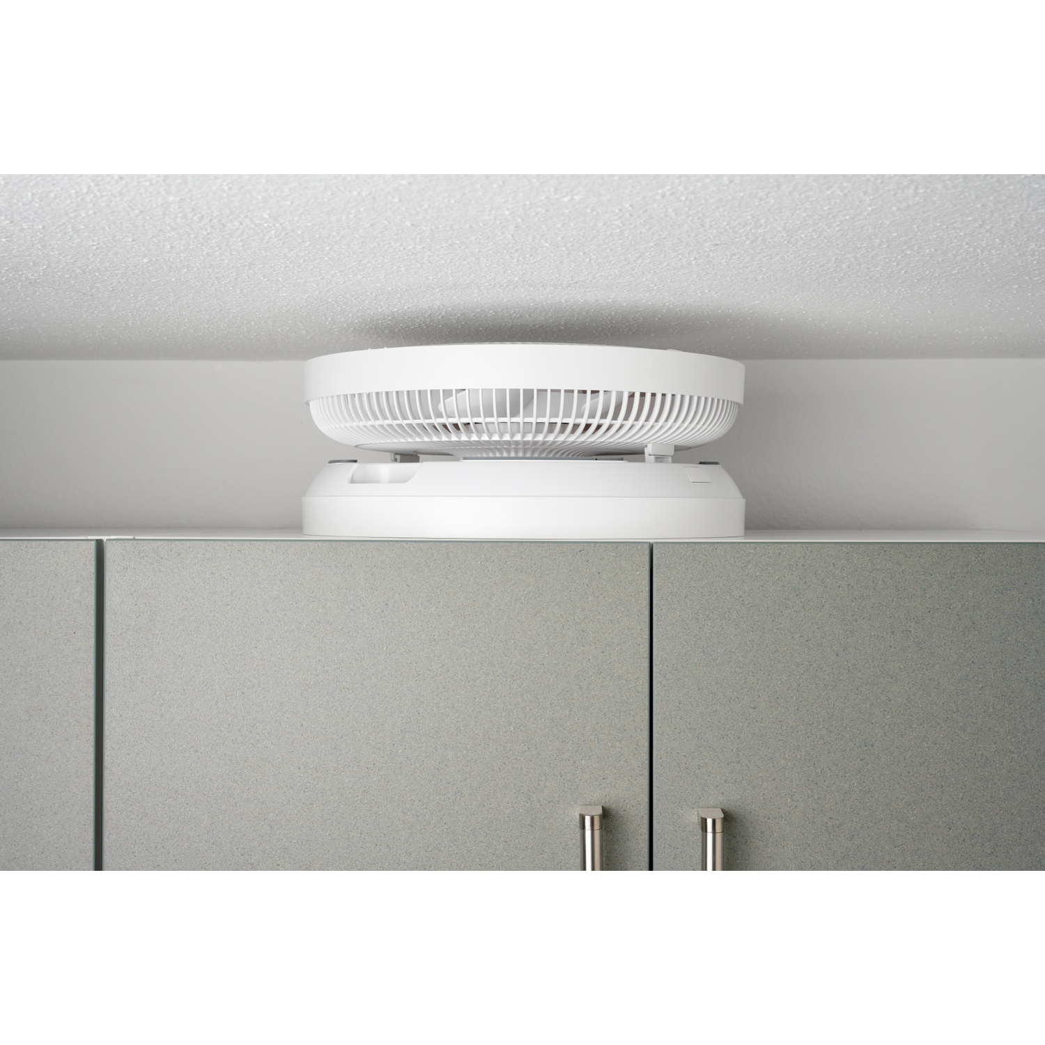 Un ventilateur blanc repose sur des portes d'armoire grises sous un plafond.
