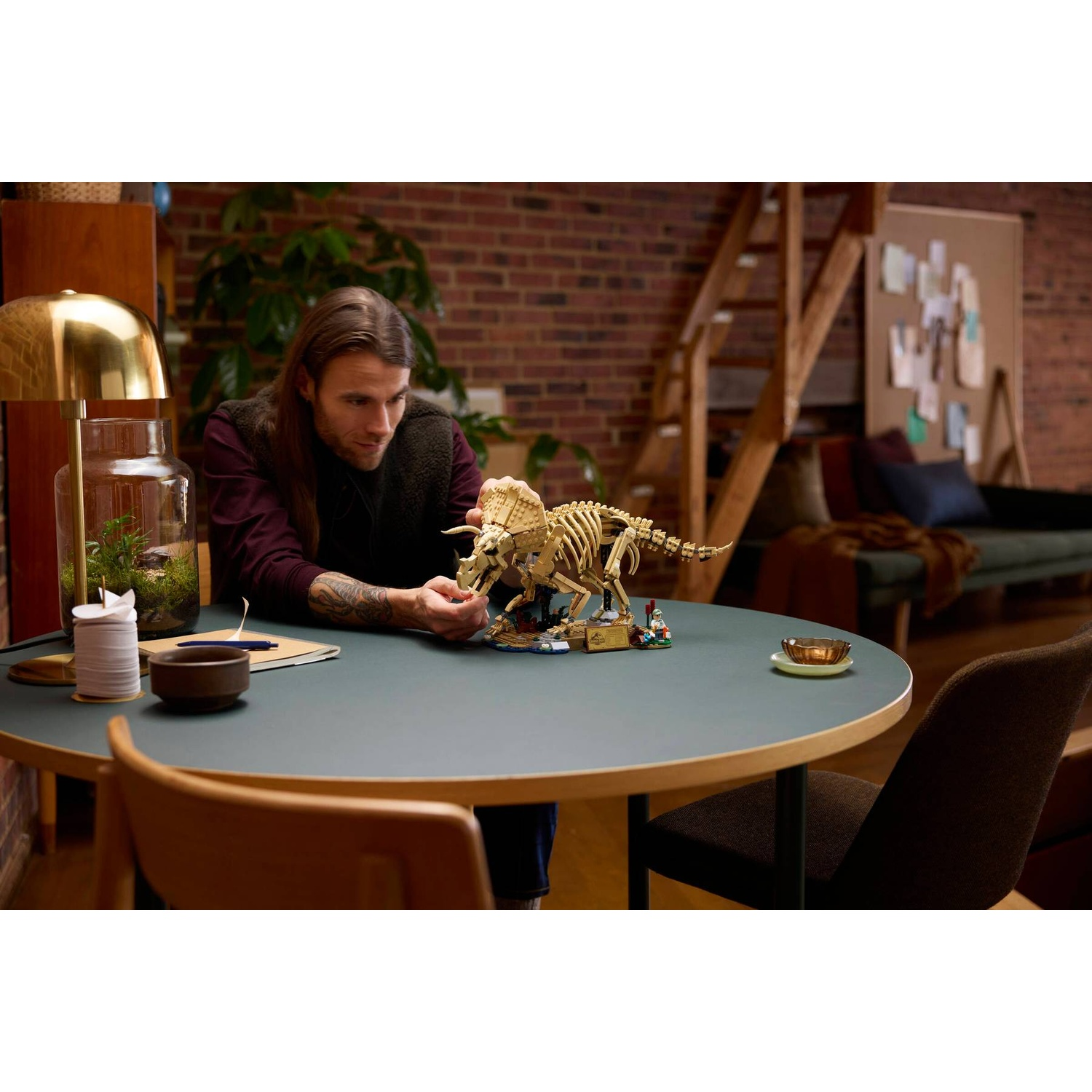 A man at a table builds a Lego dinosaur skeleton, with a lamp and terrarium nearby.