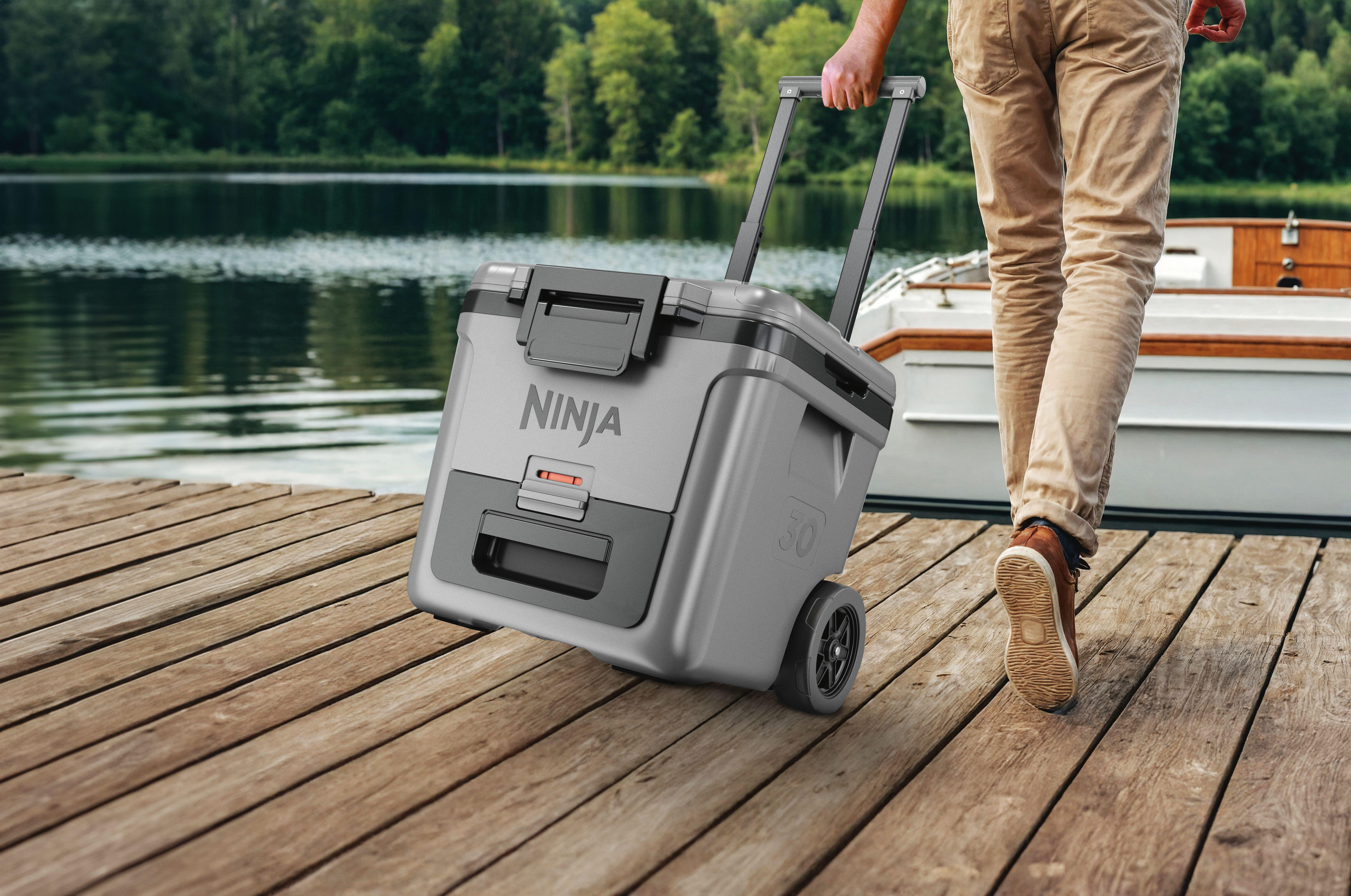 Ein Mann zieht eine graue Kühlbox auf Rollen über eine Holzplanke am See.