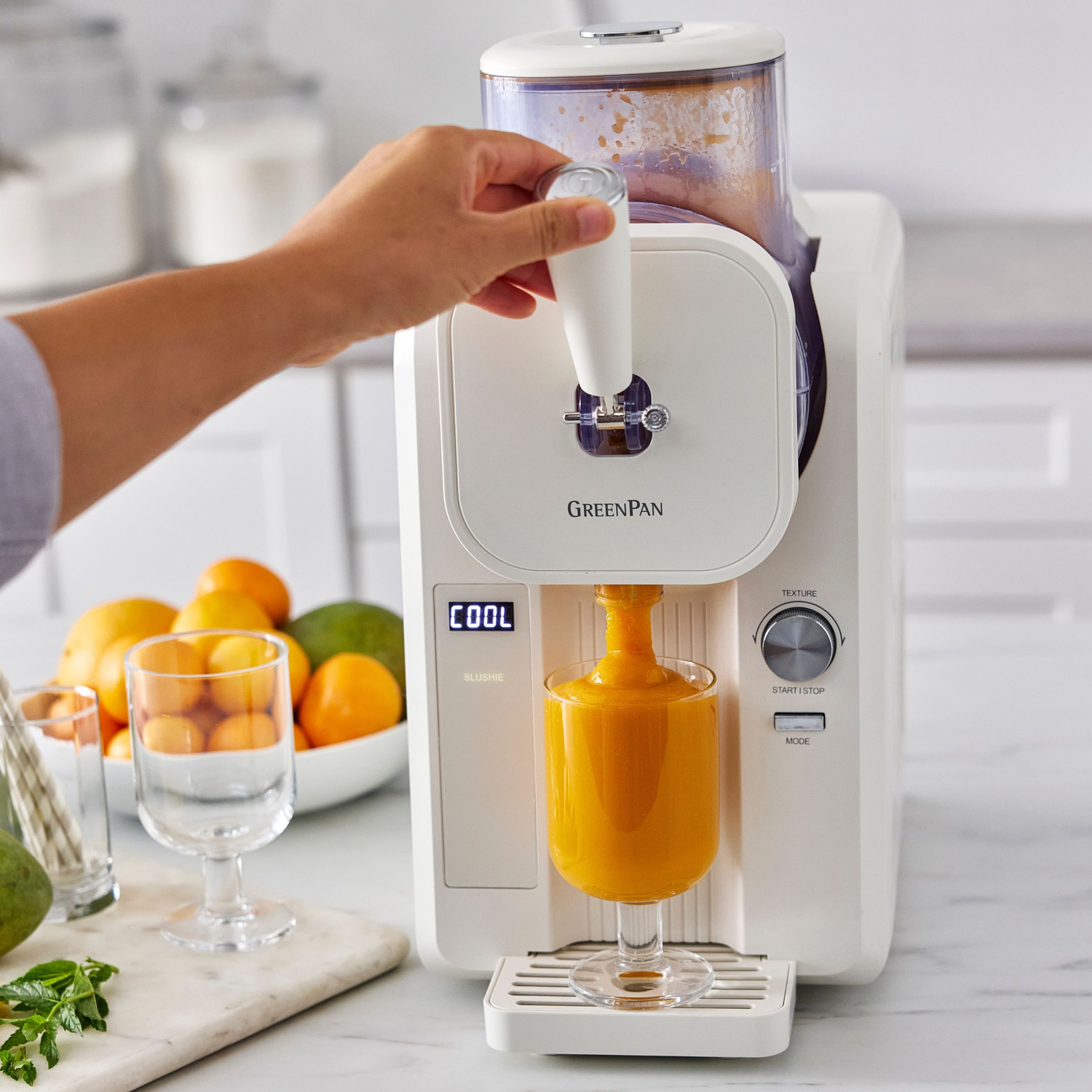 Een hand bedient een witte slushie maker die oranje slushie in een glas schenkt. Fruit op de achtergrond.