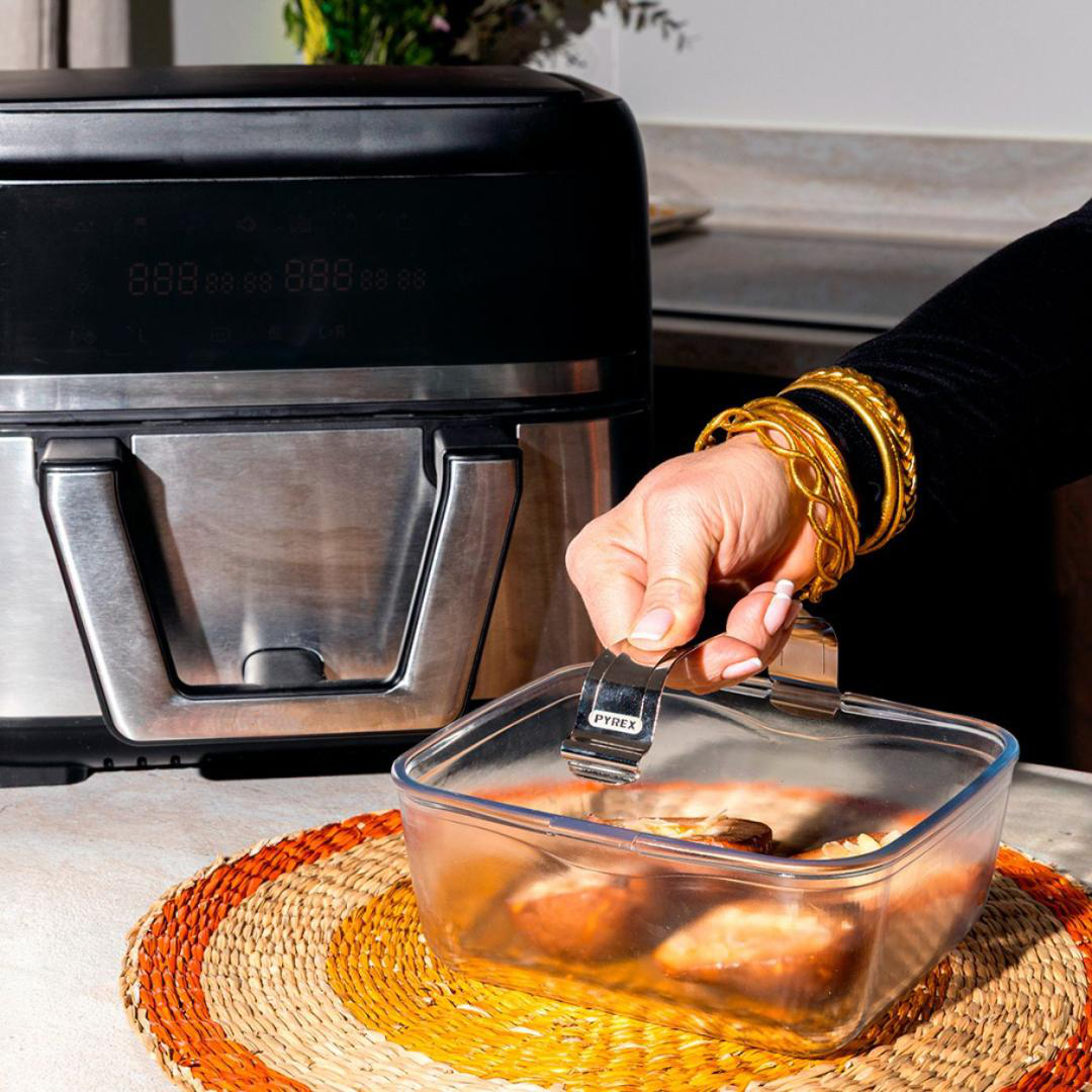 Eine Hand mit goldenen Armreifen hebt einen Pyrex-Glasbehälter mit Speisen neben einem schwarzen und silbernen Gerät an.