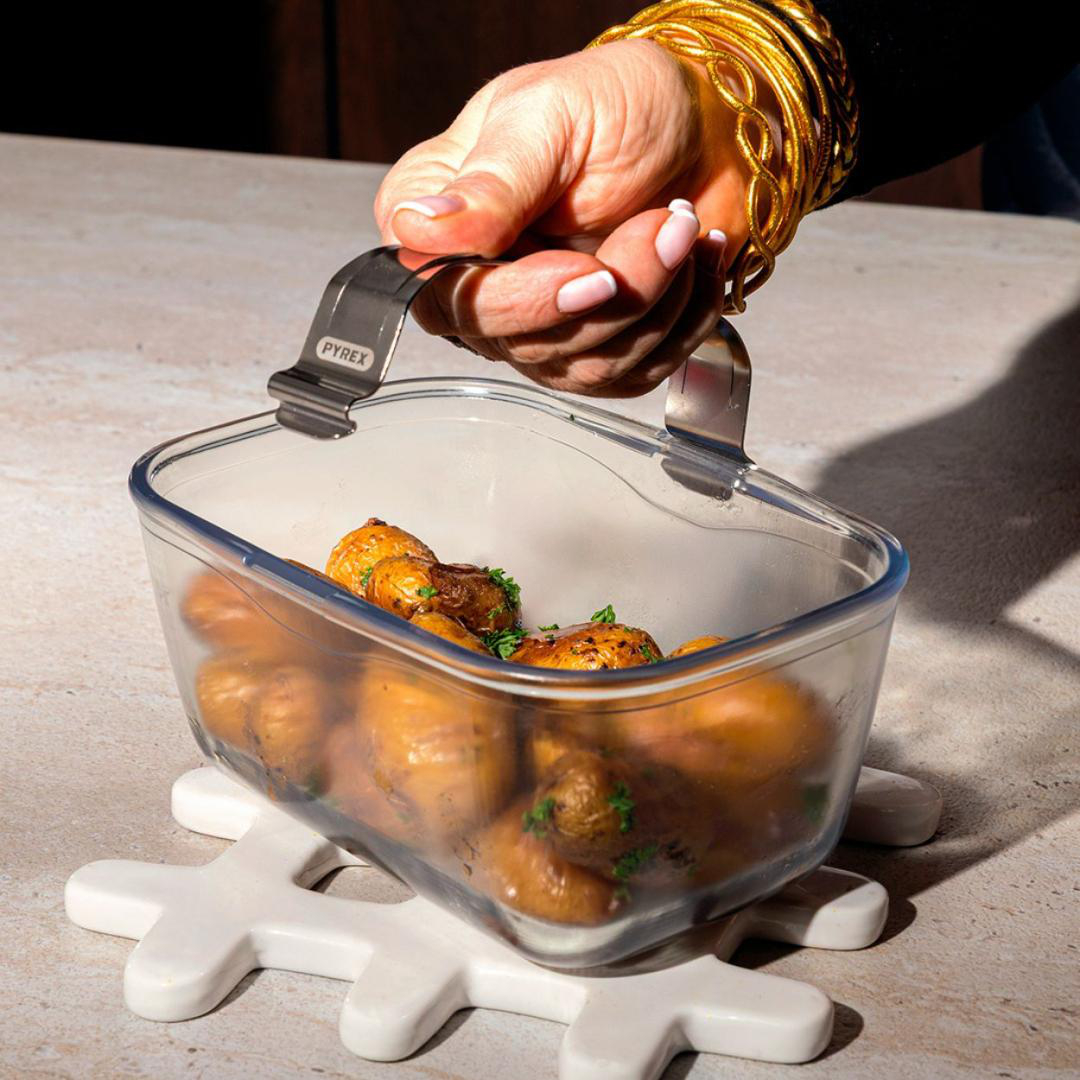 Eine Hand mit goldenen Armreifen hält eine Pyrex-Schale mit gebackenen Kartoffeln.