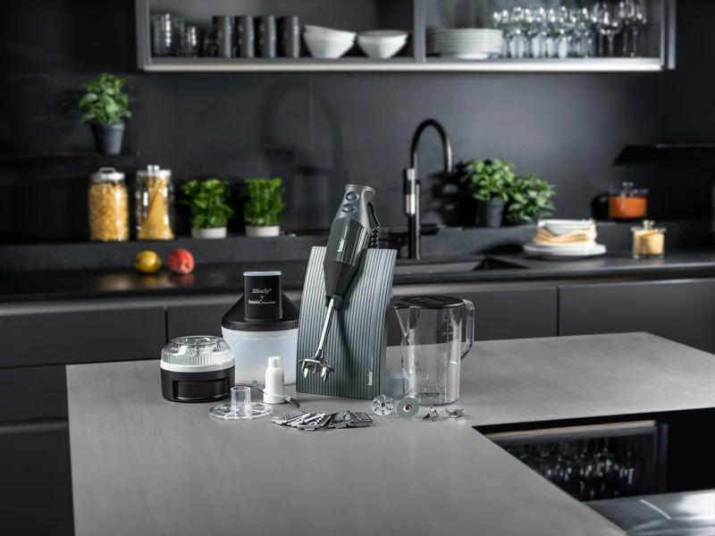 Ein Stabmixer-Set mit Zubehör steht auf einer Küchentheke mit Gläsern und Lebensmitteln im Hintergrund.