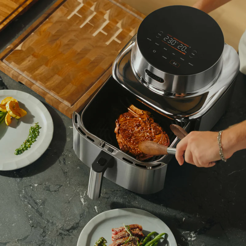 Ein Ribeye-Steak wird mit einer Zange in einen silbernen Airfryer gelegt. Daneben liegen Beilagen auf Tellern.