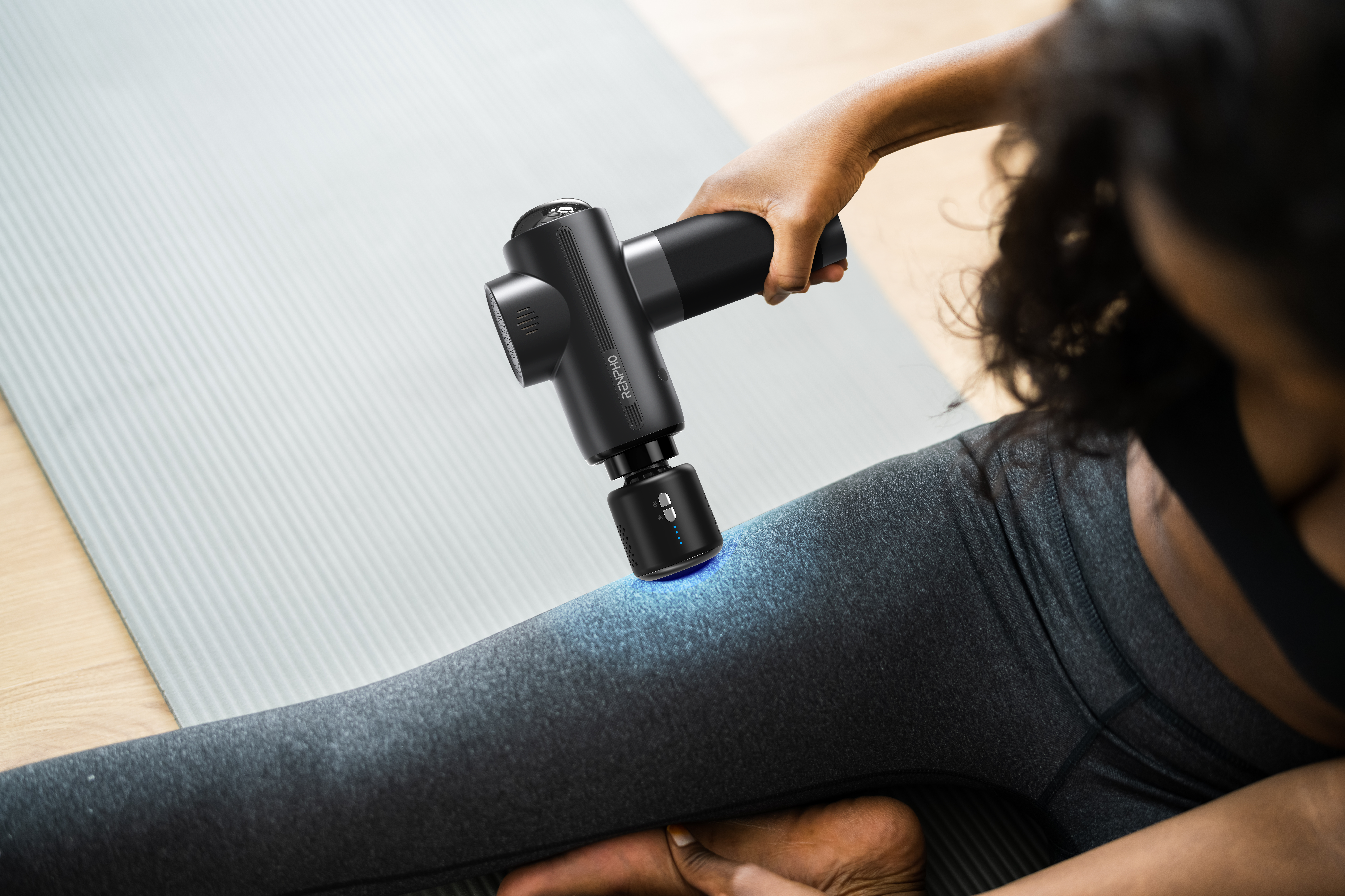 Eine Hand hält eine schwarze Massagepistole an den Oberschenkel einer Person auf einer Yogamatte.