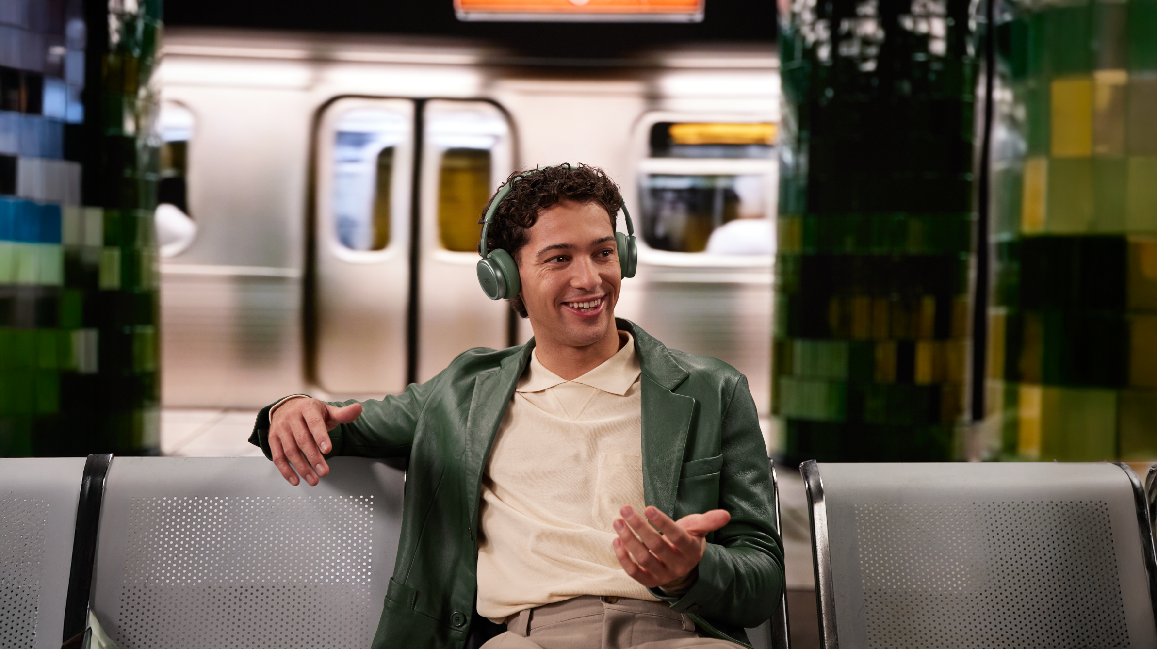 Uomo con cuffie sorride, seduto su una panchina in stazione metro con un treno dietro.