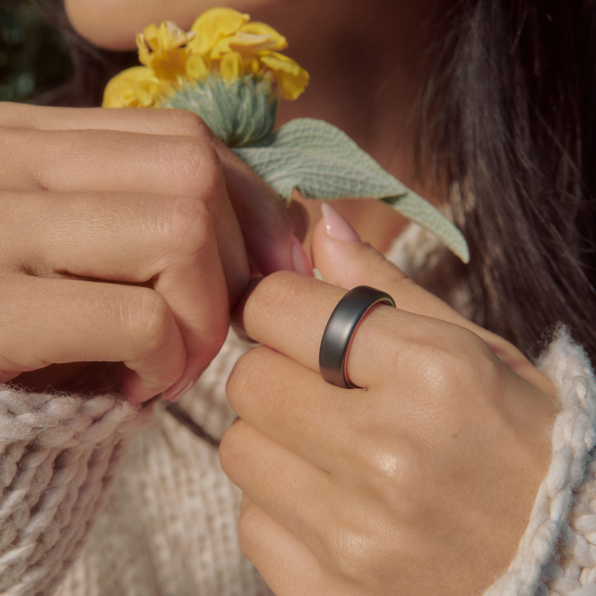 Eine Frau hält eine gelbe Blume. Sie trägt einen dunklen Ring am Finger.