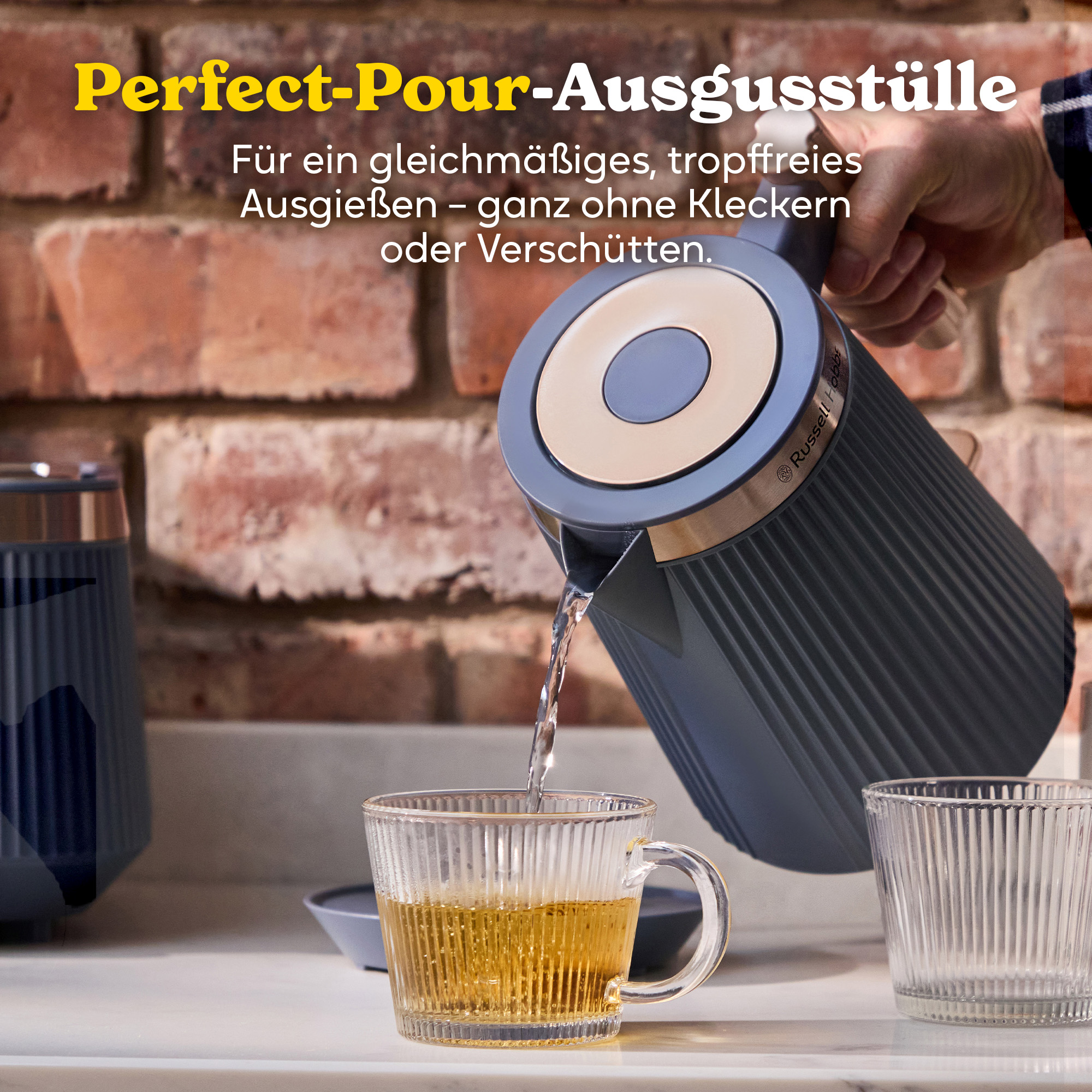 Ein dunkelblauer Wasserkocher gießt Wasser in eine Glas-Teetasse, Ziegelwand im Hintergrund.