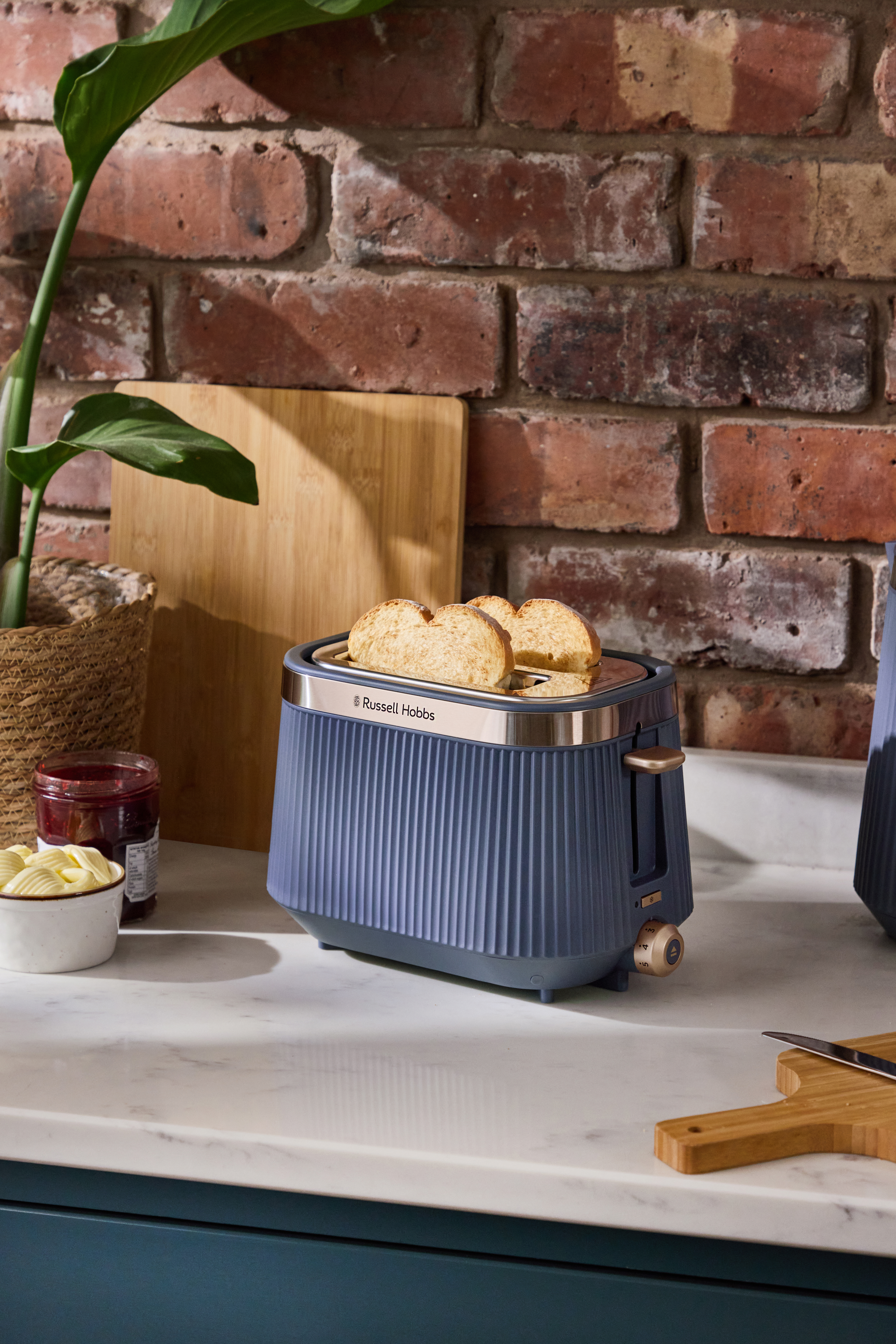 Ein blauer Russell Hobbs Toaster mit Brot steht auf einer Marmorarbeitsplatte. Marmelade und Butter sind in der Nähe.