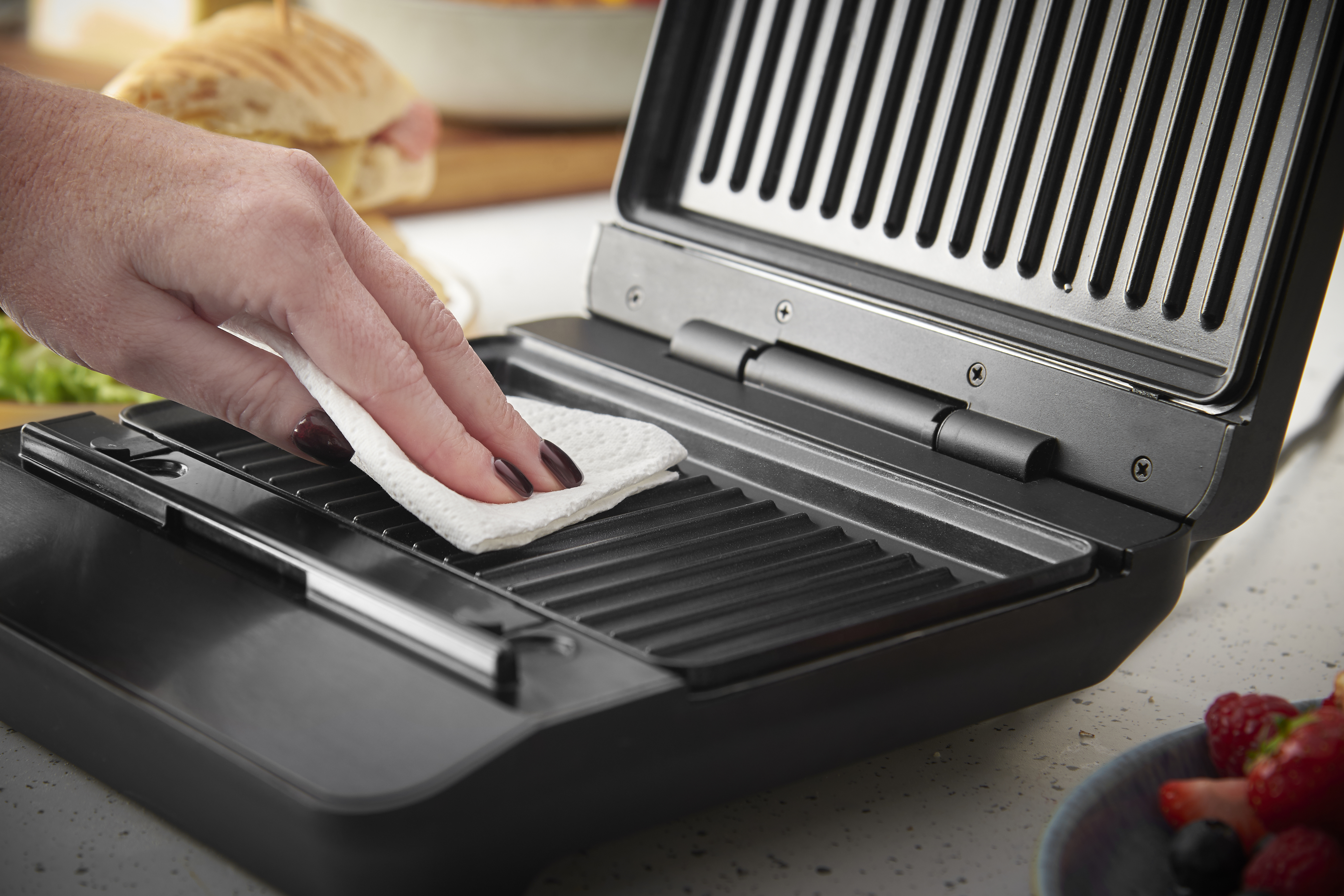 Eine Hand mit dunklem Nagellack reinigt einen Kontaktgrill mit einem weißen Tuch.