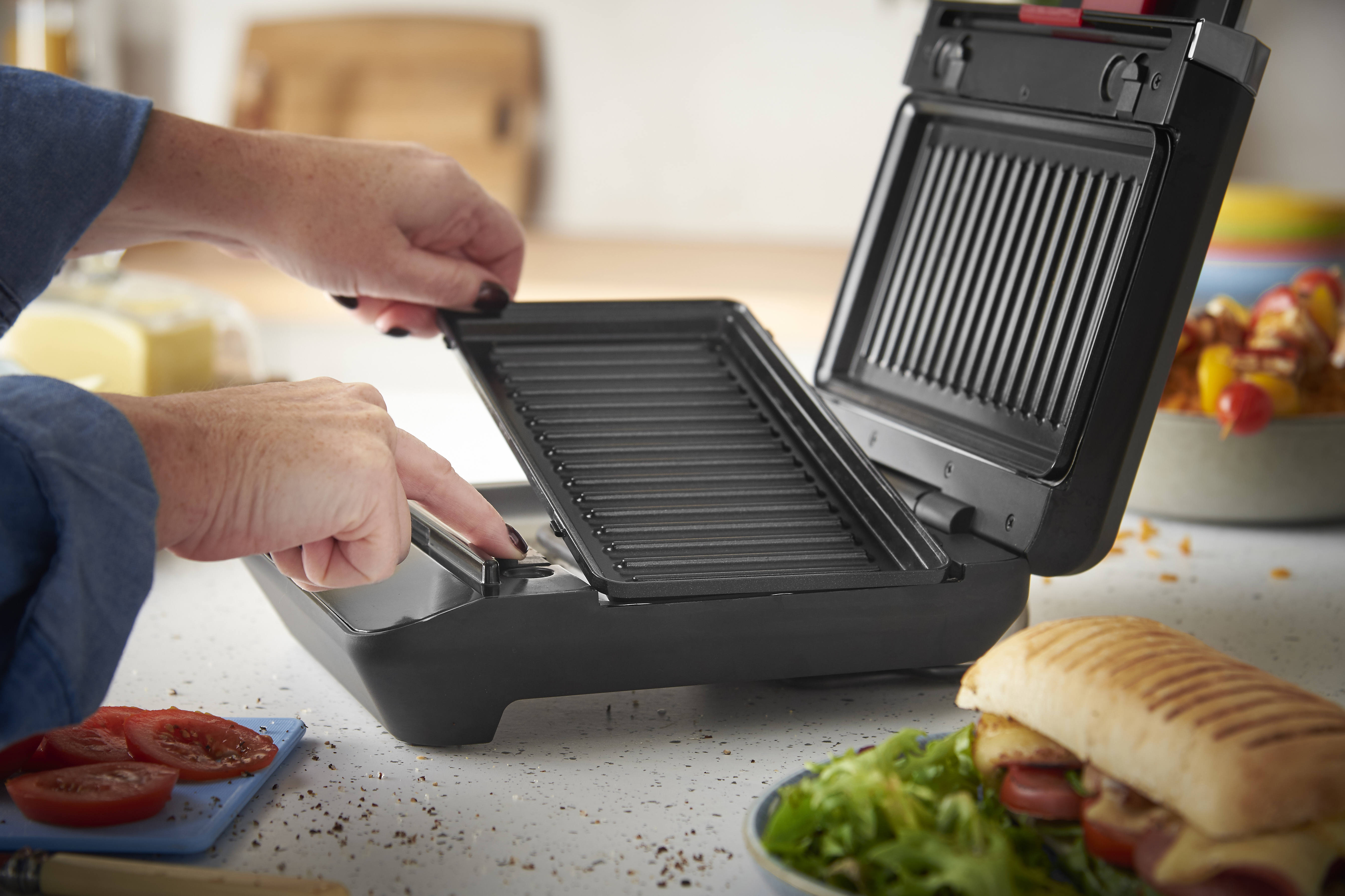 Person benutzt einen Kontaktgrill mit geöffnetem Deckel, daneben ein Sandwich.