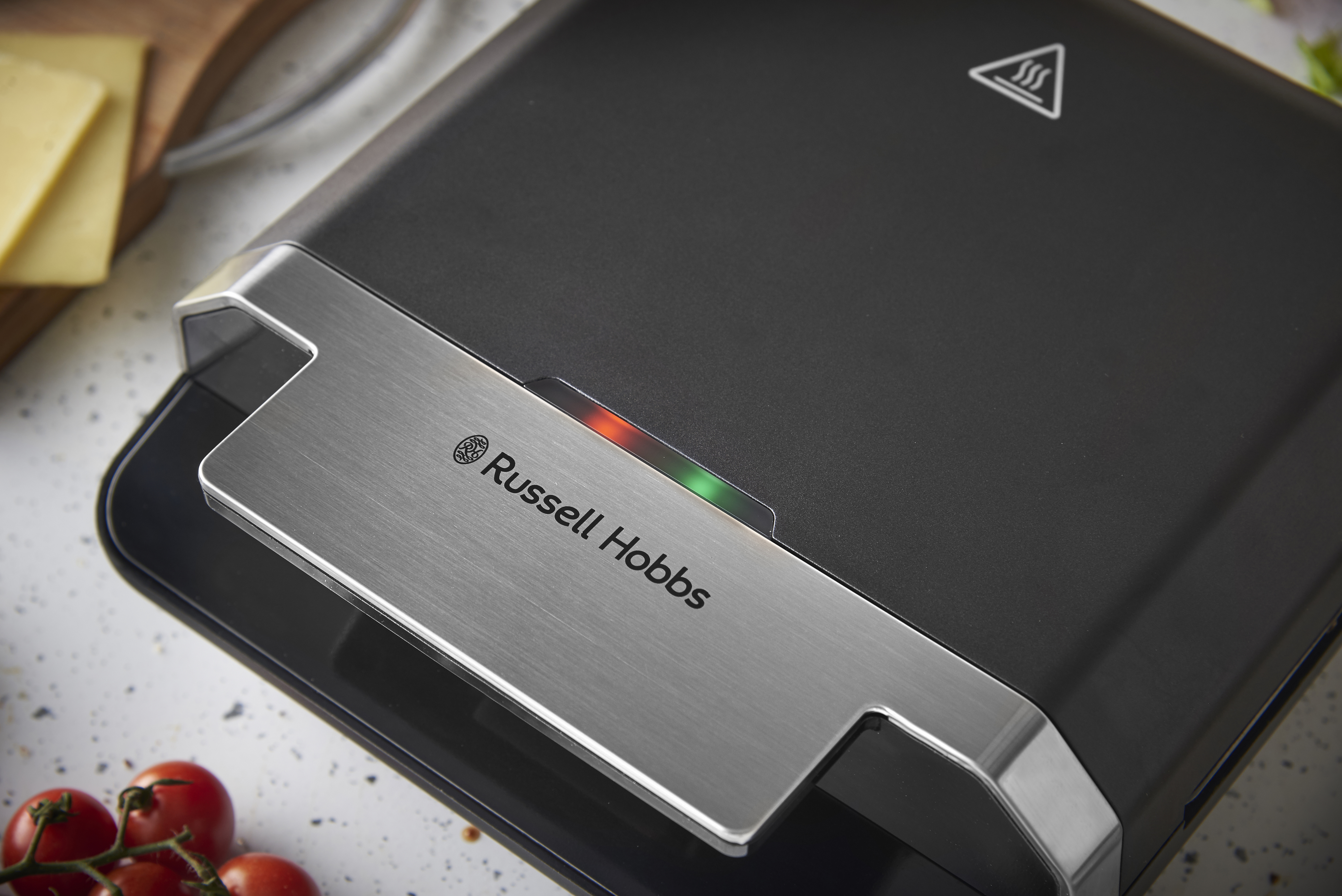 Ein schwarzer Sandwichmaker mit Russell Hobbs Logo und Lämpchen, daneben Käse und Tomaten.