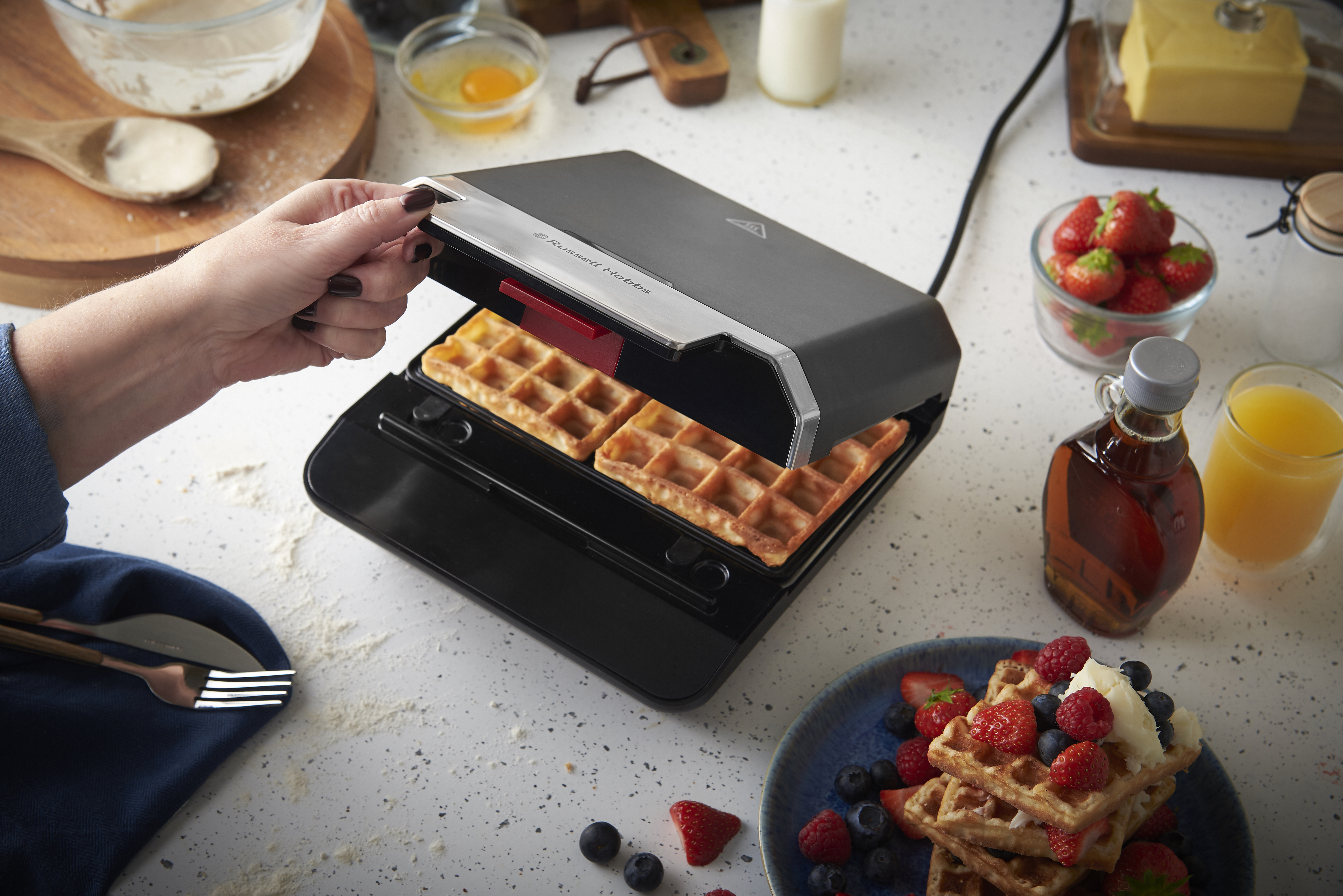 Eine Hand öffnet einen Waffelautomaten mit fertigen Waffeln, daneben Beeren und Saft.